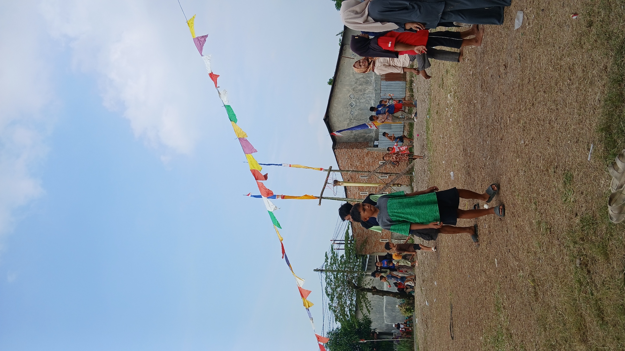 The image depicts a scene outdoors in Klambir Lima Kebun, Indonesia, under a bright blue sky with scattered clouds. A string of colorful triangular flags stretches across the frame, strung between poles and extending into the distance. In the foreground, a young boy wearing a green t-shirt and a dark skirt is looking towards the viewer. Behind him, other individuals, including children and adults, are visible, some standing and others in motion.
Several buildings are present in the background, including a brick structure with a gray roof and another building with a corrugated metal wall. The ground is covered with dry grass and some debris. The overall atmosphere suggests a community gathering or event, possibly a celebration or festival, given the presence of the flags and the crowd. The area appears to be a rural or semi-urban setting.