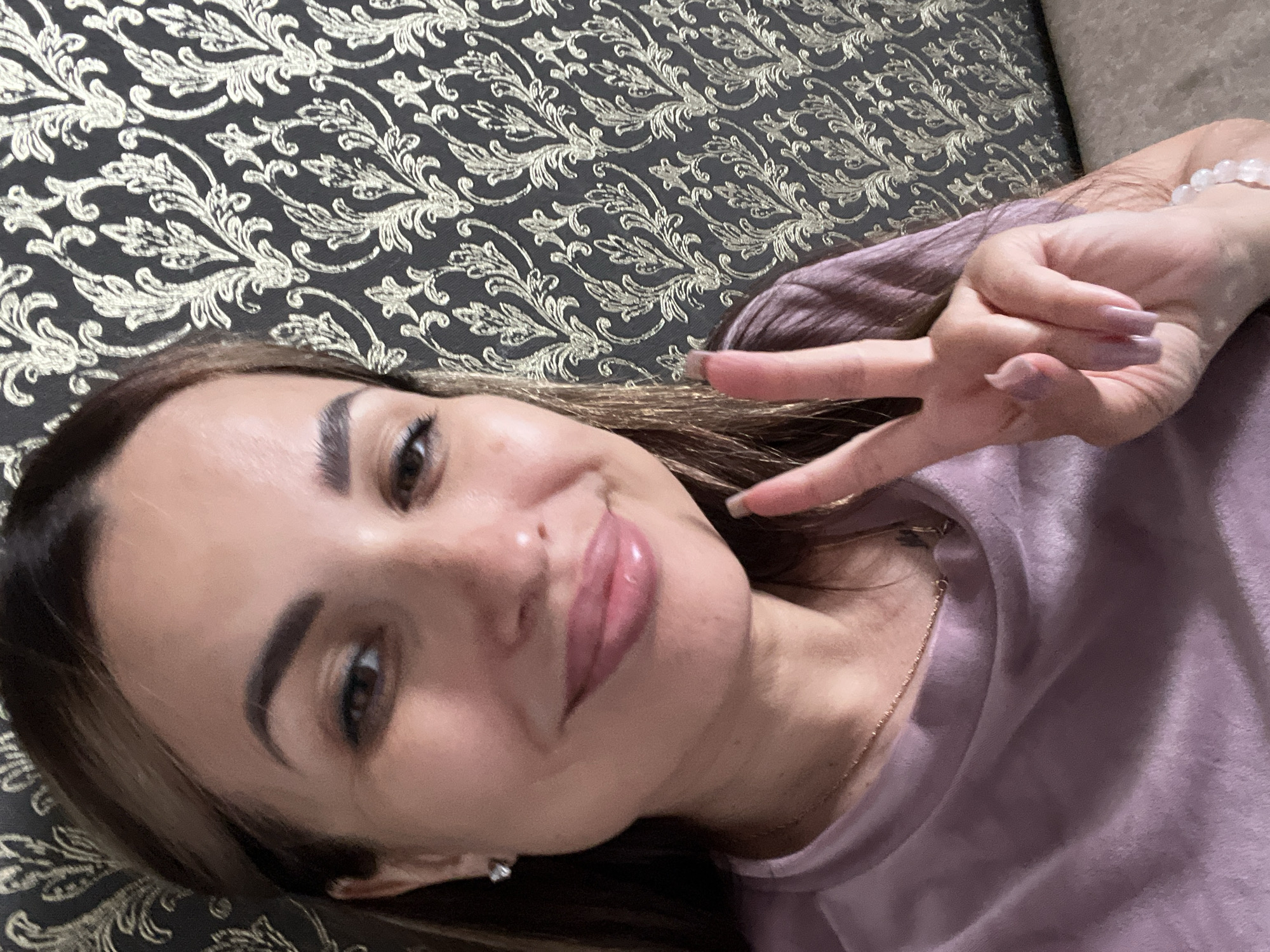 A woman with long brown hair, wearing a purple shirt and a necklace, smiles as she holds up two fingers in a peace sign. She has on light pink lipstick and is wearing diamond stud earrings. The background is a dark gray wallpaper with a gold ornate pattern. The lighting is soft and natural.