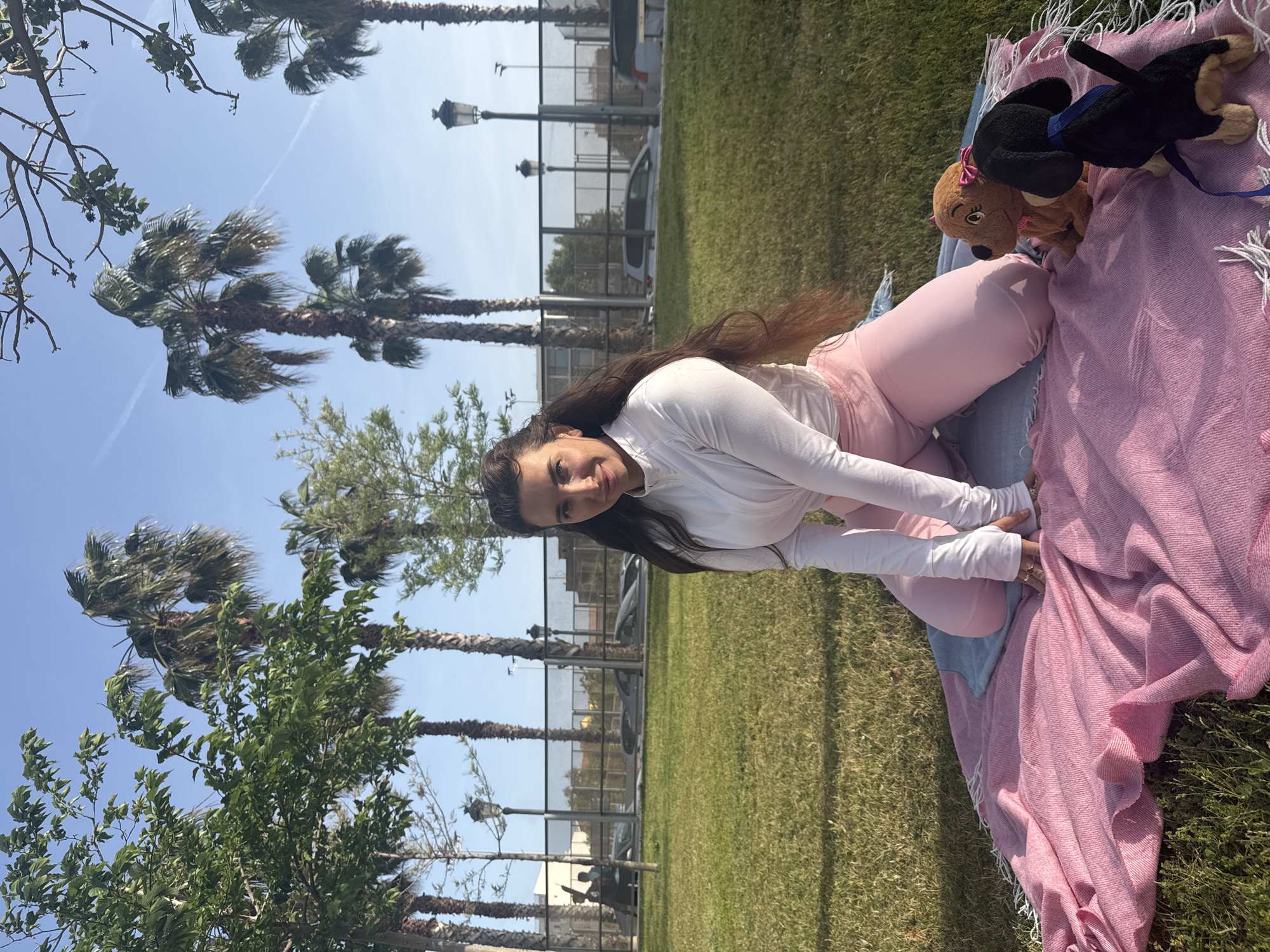 An outdoor photograph, correctly oriented, depicts a young woman relaxing in a sunny park or green space, likely in València, Spain. The woman, with long, dark brown hair, is wearing a white long-sleeved top and pink leggings. She is sitting or kneeling on the vibrant green grass, smiling warmly towards the camera, with her hands gently resting on a pink woven blanket with white fringe that is spread beside her.
On the blanket, two plush dog toys are visible: one light brown with a pink bow on its head, and another black toy dog with a blue leash or collar.
The background features several tall palm trees standing against a clear, bright blue sky, with a visible white jet contrail. Below the palms, a black metal fence separates the grassy area from what appears to be a road or parking lot, where parts of parked cars, including a white van and a dark vehicle, can be seen. Ornate streetlights add a decorative touch to the urban landscape. Lush green foliage from other trees and shrubs frames the scene on the left, contributing to the serene park atmosphere. The bright daylight and the woman's relaxed demeanor suggest a pleasant, leisurely moment. No visible text is present in the image.