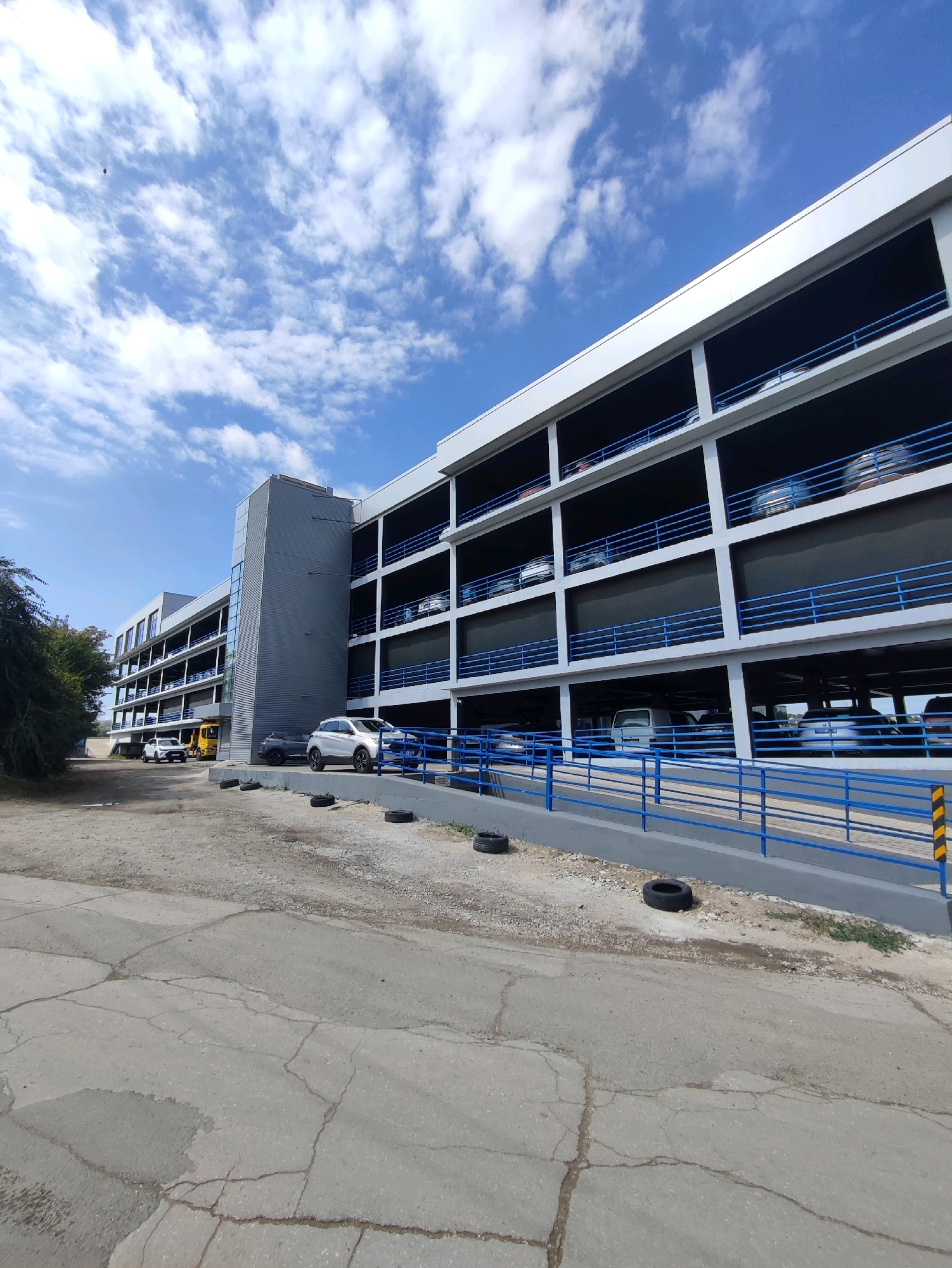 The image depicts a modern, multi-level parking garage under a bright blue sky with scattered white clouds. The garage has a sleek, contemporary design with concrete floors and white exteriors, accented by blue railings on each level. Cars are visible parked on various levels, suggesting it is in use. The ground in front of the garage is a cracked asphalt or concrete surface, with some gravel and scattered tires, indicating a functional but perhaps less manicured area. To the left of the garage, there is a patch of green trees and foliage. The overall scene is one of urban infrastructure, with the parking structure being the dominant element. The location is identified as Chișinău, Moldova.
