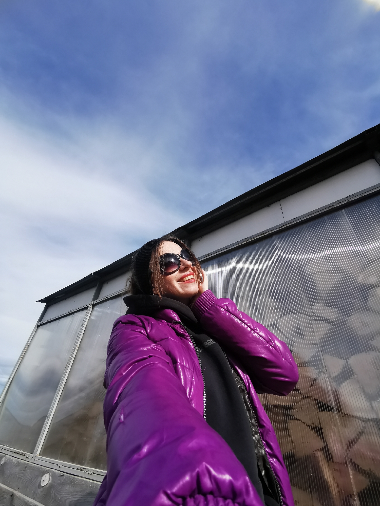 A woman wearing a black beanie, sunglasses, a black hoodie, and a shiny purple puffer jacket stands outside a building. The building has white siding at the top and a clear ribbed plastic facade below, suggesting it might be a greenhouse or similar structure. The sky above is blue with scattered clouds. The exact location and city are not discernible from the image.
