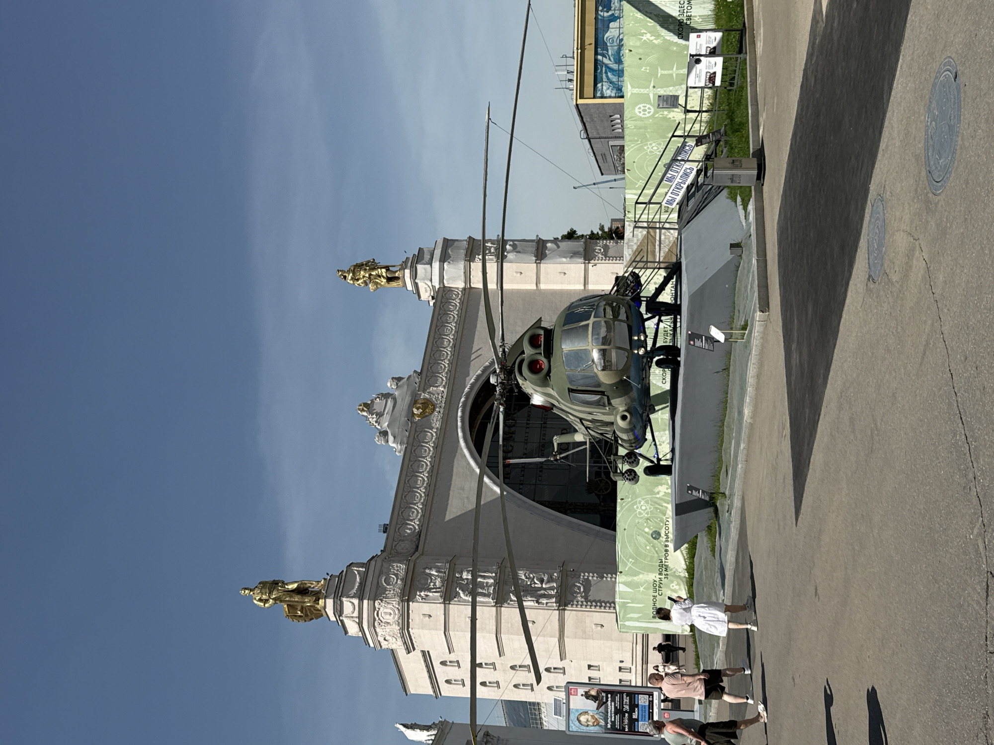 The image shows a helicopter on display in front of a grand, ornate building with golden statues. The building's architecture is classical, with detailed carvings and arches. The helicopter is an olive-green military-style aircraft. Several people are visible, some walking by and others seemingly observing the exhibit. A large banner or sign is near the helicopter, featuring text in Russian. The location appears to be a public space, likely a park or exposition ground in Moscow, Russia, based on the architecture and signage. The day appears bright and sunny, judging from the clear blue sky and shadows.