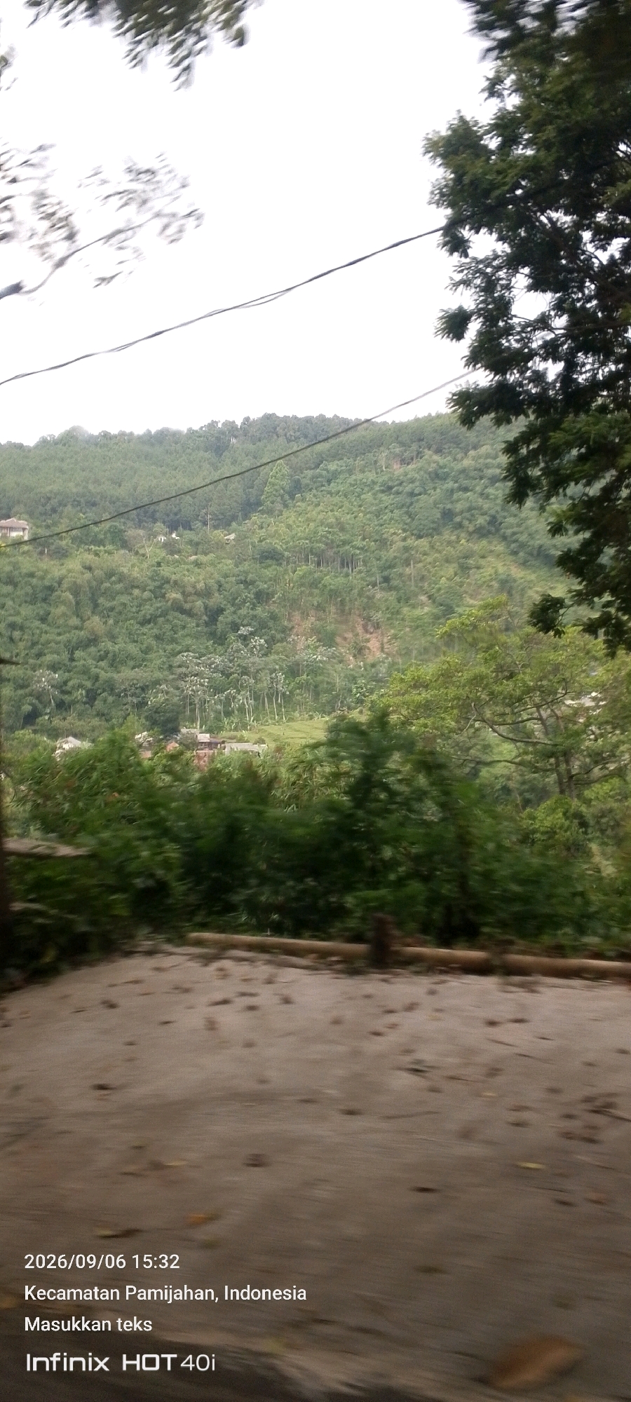 The image shows a hillside covered in dense green foliage, with some scattered buildings visible among the trees. In the foreground, there is a dirt or gravel path with some fallen leaves. The sky is overcast and white. Text overlay on the image indicates the date "2026/09/06 15:32", the location "Kecamatan Pamijahan, Indonesia", and the device "Infinix HOT 40i".