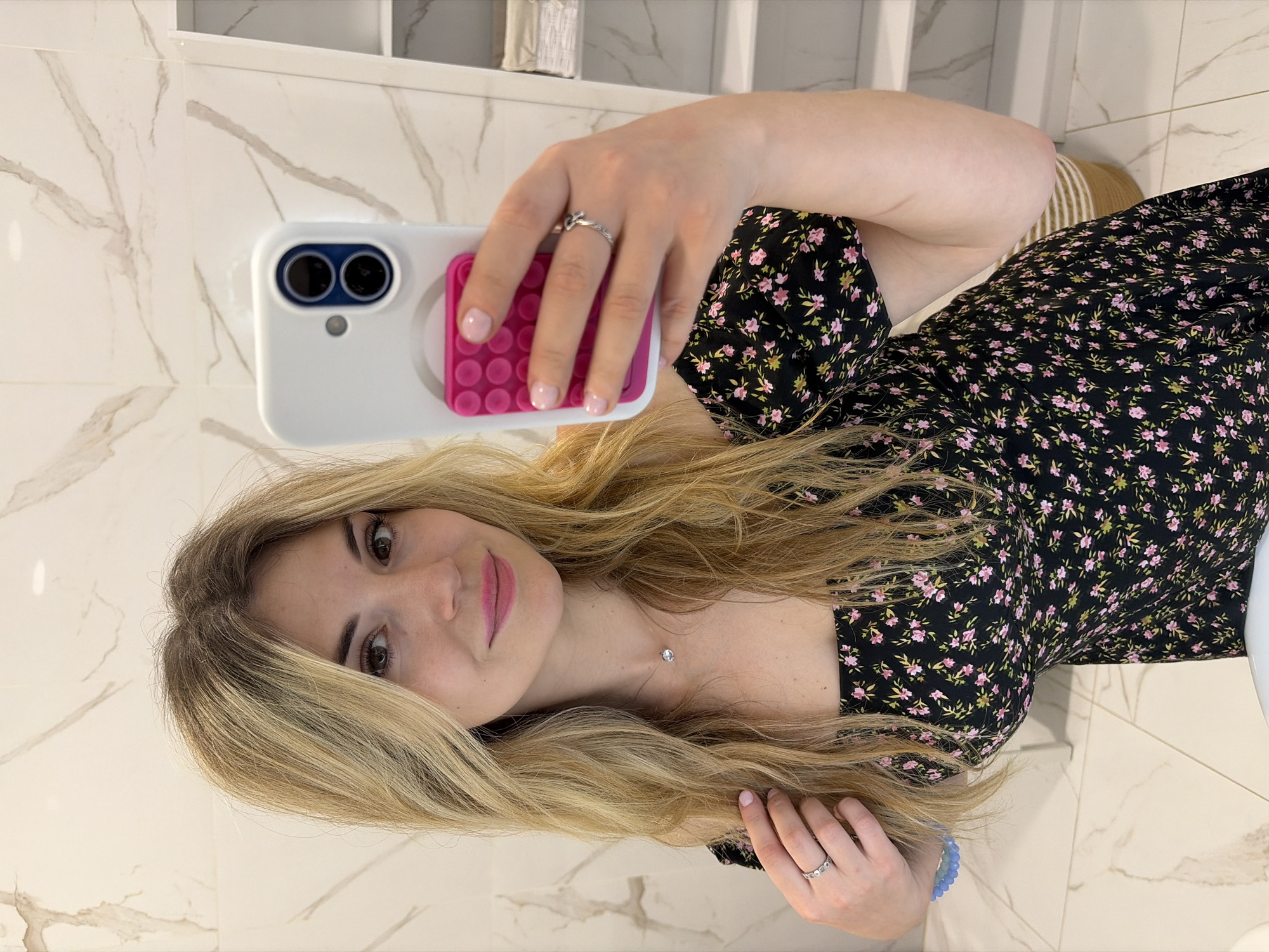 A woman with long blonde hair, wearing a black floral dress and jewelry (rings and necklace), is taking a selfie with a white smartphone. She is indoors, in what appears to be a bathroom, judging by the marble-patterned wall and floor tiles and bathroom fixtures visible in the background. The location is the city of Budva, Montenegro.