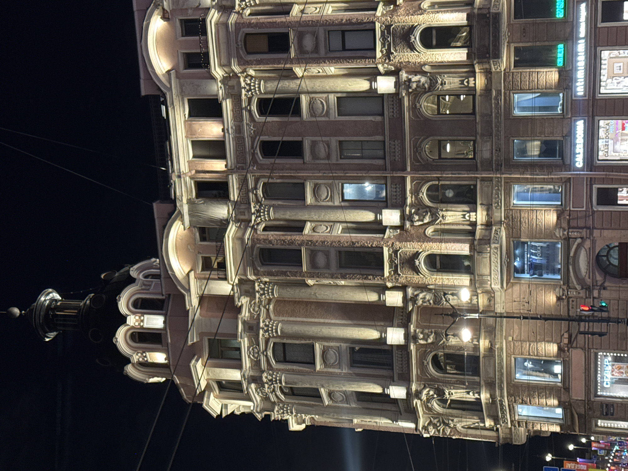 The image displays a grand, ornate building illuminated at night in an urban setting, consistent with Saint Petersburg, Russia. The multi-story structure features intricate classical architecture, including decorative columns, detailed cornices, carved reliefs, and a mix of arched and rectangular windows. A prominent cupola or dome-like element crowns the building at the top.
The facade, rendered in light tones, is dramatically lit by artificial lights, highlighting its elaborate details against the dark night sky. Many windows are dark, while others glow with internal light, revealing glimpses of interior spaces or shop displays. Overhead power lines crisscross the scene.
On the lower levels, several retail shops are visible. Clear signage for "CALZEDONIA" and "INTIMISSIMI" is prominently displayed, with the latter accompanied by green digital numbers such as "627 628." Further down, partial Cyrillic text, "CBEP" (likely Sber), can be seen in a shop window. Streetlights and a traffic light showing a green signal are also present. The absence of people emphasizes the architectural subject.