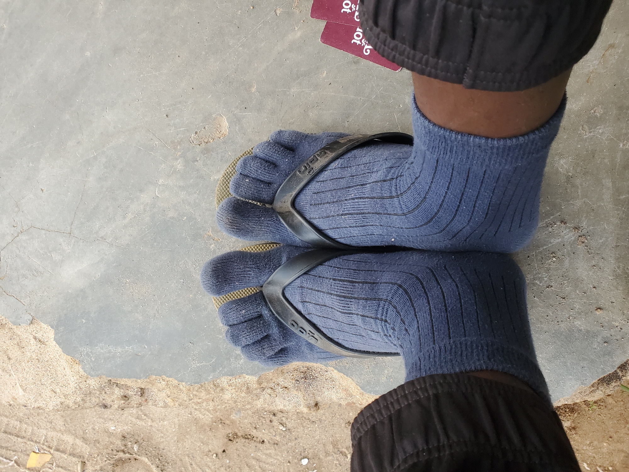 A close-up, eye-level shot shows a person's feet wearing blue toe socks and flip-flops. The flip-flops have black straps that separate each toe. The socks are ribbed and cover the ankles. The feet are resting on a concrete surface next to a patch of sandy dirt. Two red cards with white lettering are visible to the right of the feet. The scene appears to be outdoors, with natural light illuminating the area. The specific location in Bukuru, Nigeria is not discernible from the visual information provided.