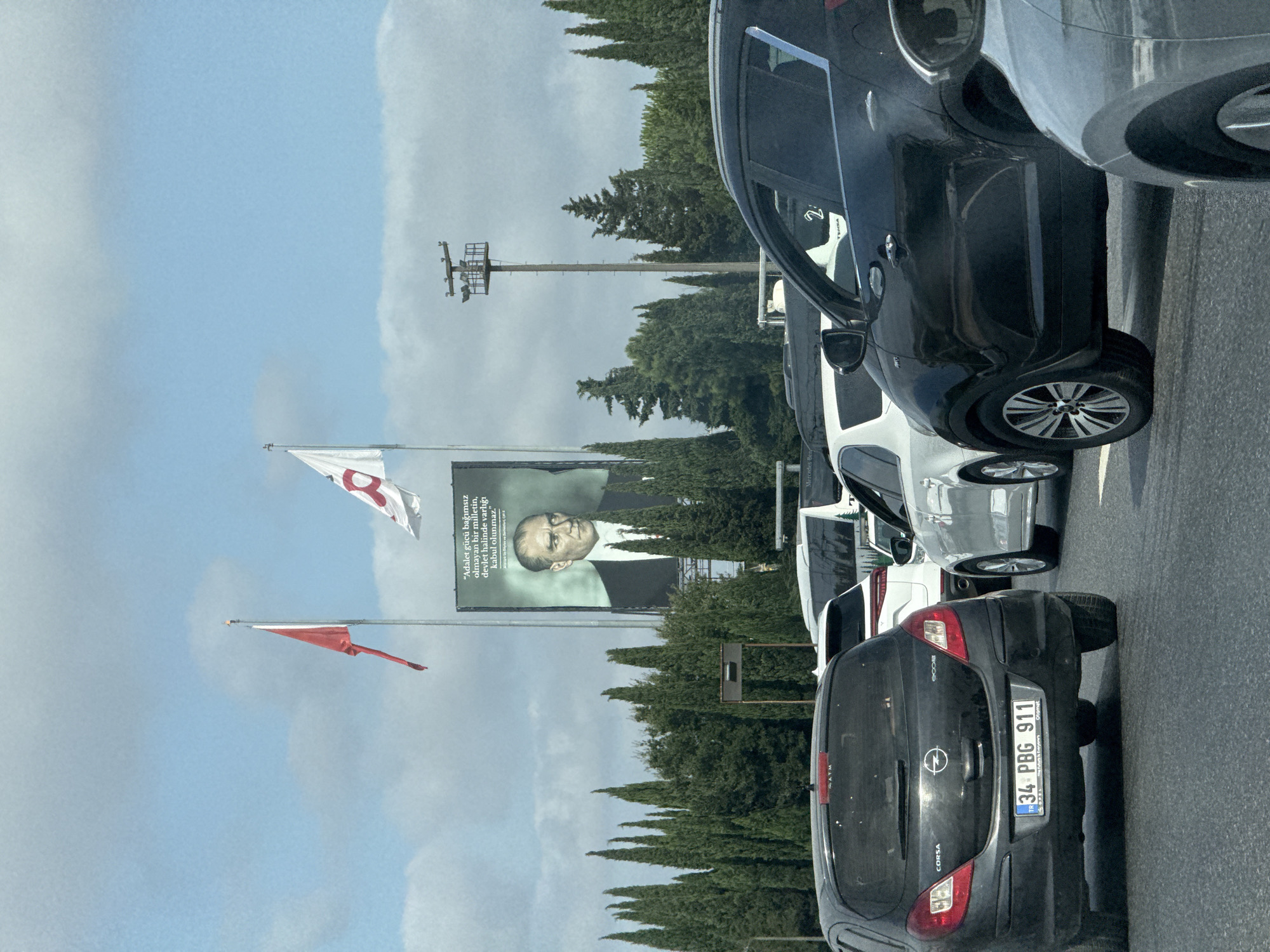 The image shows a view from a car in traffic on a highway. In the background, a large billboard displays a portrait of Mustafa Kemal Atatürk, the founder of the Republic of Turkey, with text in Turkish. Two flags are visible on poles in front of the billboard. The foreground features several cars, including a black Opel Corsa with a Turkish license plate, a white SUV, and a black SUV. Tall cypress trees line the median of the highway, and a light pole with several lights is visible. The sky is partly cloudy. The scene appears to be in a city environment with a highway setting.