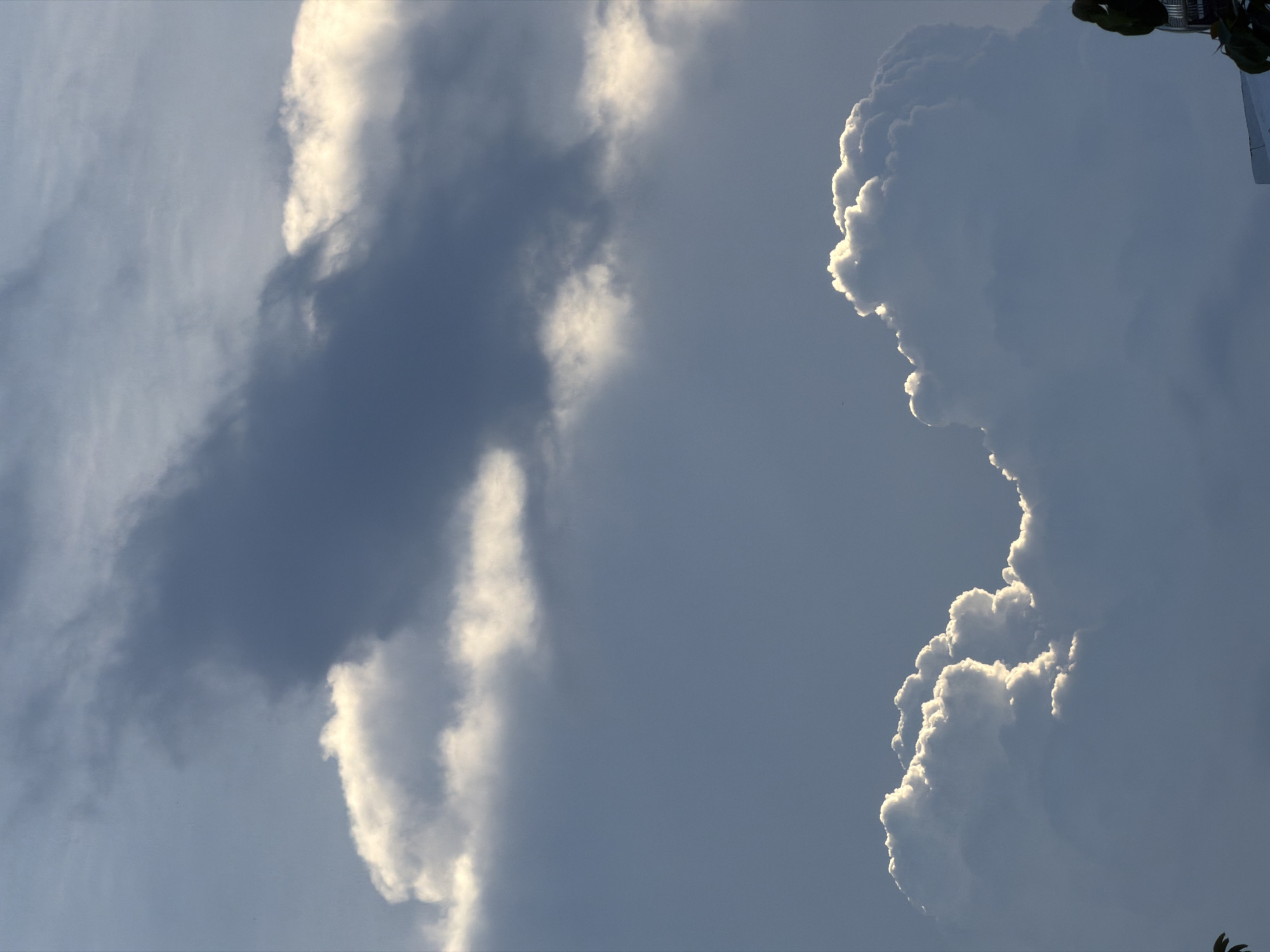 The image depicts a daytime sky with cumulus clouds. The image is taken in the city of Nha Trang Ward, Vietnam, as indicated by the metadata. One cloud formation on the right side of the image is dense and puffy, with a bright, sunlit edge. To the left is a darker, less defined cloud formation. The overall sky is a light blue-grey color. The bottom edge of the photo features the silhouette of foliage.