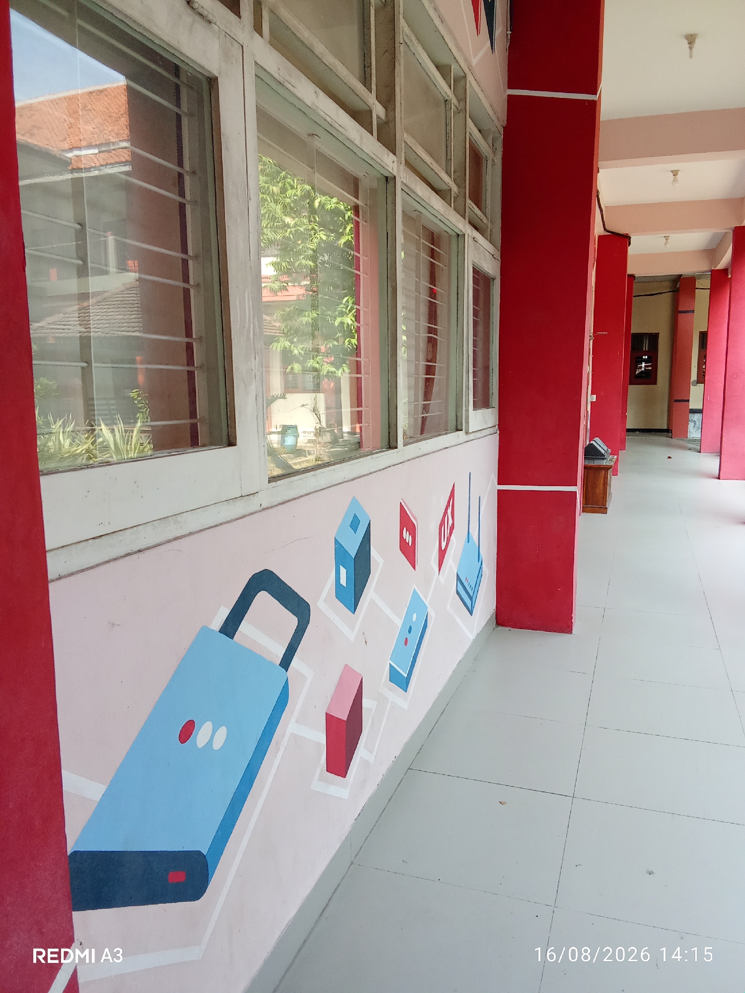 This image depicts a side view of a school hallway with a mural painted on the wall. The mural features stylized illustrations of a lock and various electronic devices. The hallway has red pillars and white window frames. The floor is tiled and appears to be a light color. The overall impression is that of a public building, likely a school, located in Surakarta, Indonesia. The date and time stamp "16/08/2026 14:15" is visible in the lower right corner.