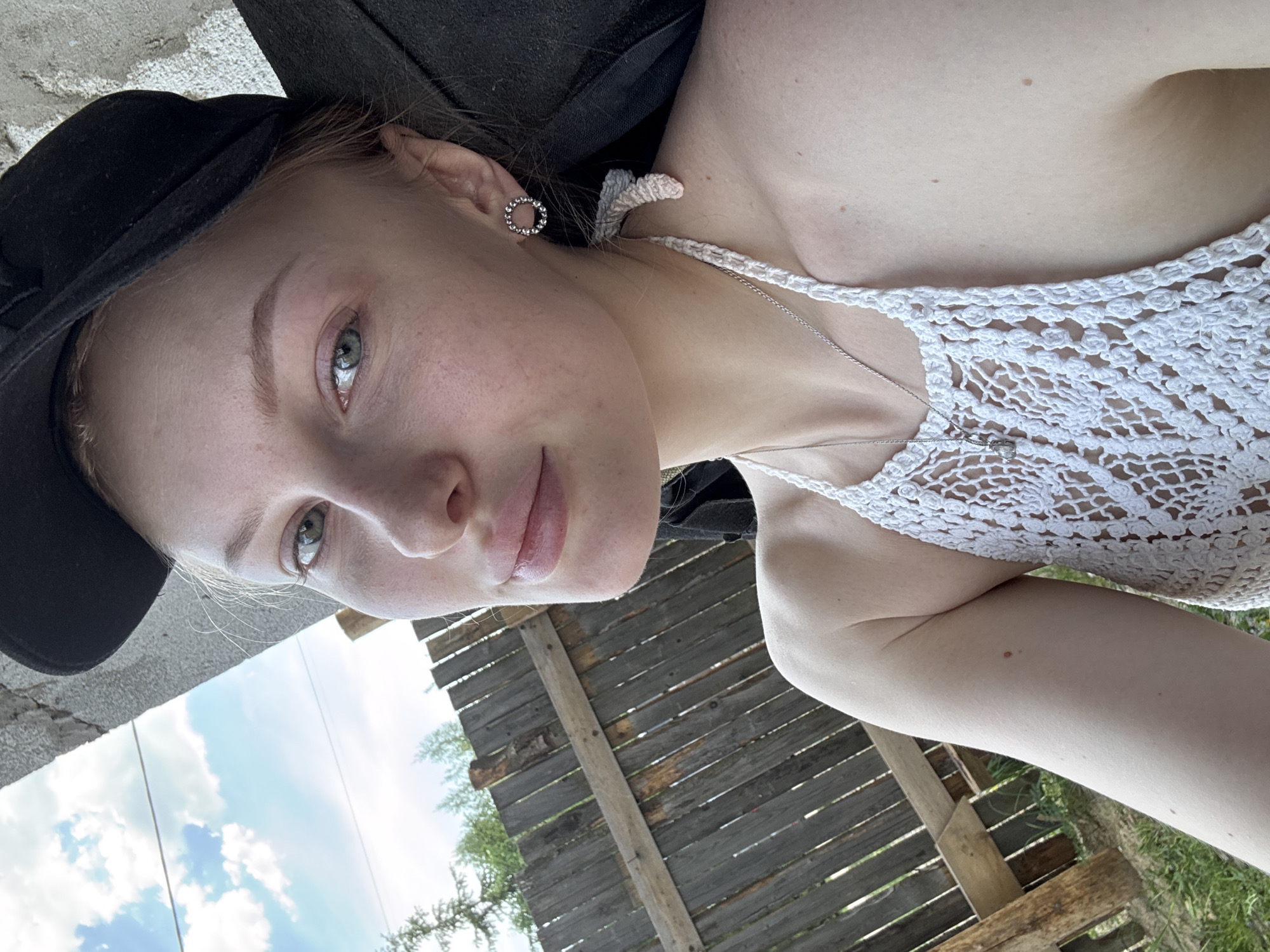 A close-up selfie photograph shows a fair-skinned person with light blonde hair, light eyes, and light pink lips. The person is wearing a black hat and a white crocheted top with thin straps. A silver necklace with a pendant is visible. A silver earring is in the left ear. The background includes a wooden fence, green foliage, blue sky, and white clouds. There is no information in the image to identify this location in Perm, Russia.