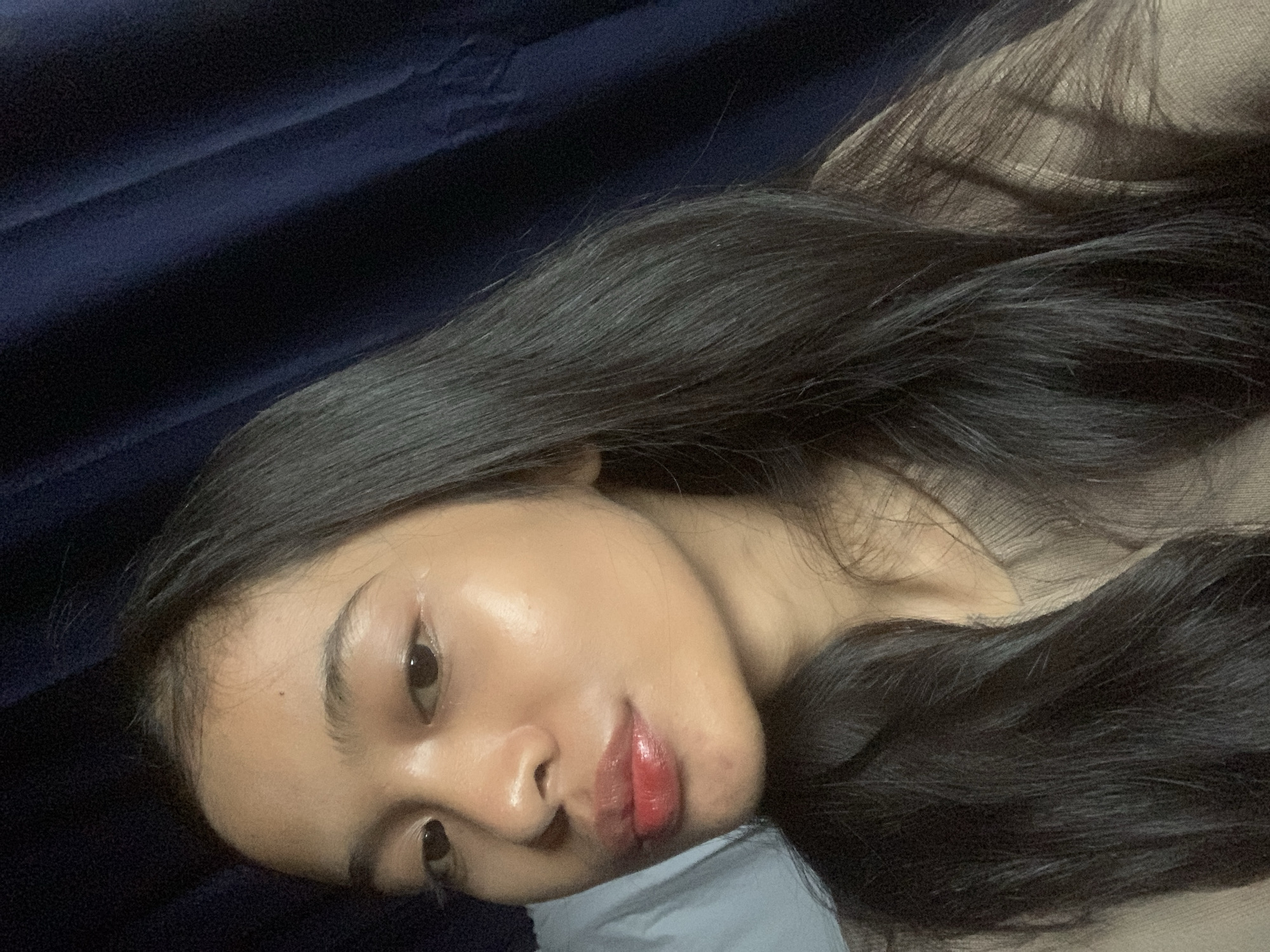 The image shows a close-up, overhead shot of a young woman lying down, with her head tilted to the left. She has long, dark hair that is spread out around her. Her face is turned towards the camera, with her eyes open and looking directly at the viewer. She is wearing light-colored clothing. The background is dark and appears to be a fabric surface. A pillow is visible beneath her head, suggesting she is in a relaxed or resting position. The lighting is soft, highlighting the sheen on her skin and lips, which are painted with a glossy red lipstick. There are no discernible text elements in the image. The context provided suggests the location is Sungai Meriam, Indonesia.