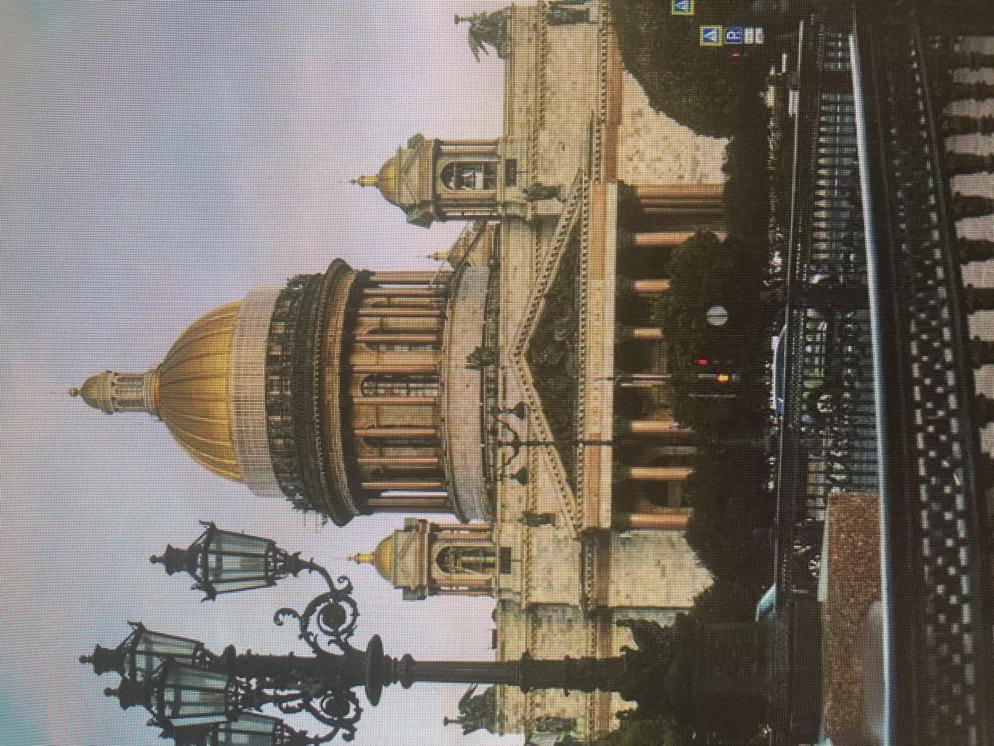 The image shows a wide shot of Saint Isaac's Cathedral in Saint Petersburg, Russia. The grand golden dome of the cathedral dominates the background, with smaller domes and bell towers flanking it. The facade of the cathedral is made of light-colored stone, with numerous columns and architectural details. In the foreground, a decorative black lamppost with multiple lights stands to the left. A dark metal fence runs across the middle ground, behind which trees and the lower portions of the cathedral are visible. A street with cars and traffic lights is suggested by elements in the middle and right ground. The sky is overcast, casting a soft light on the scene.