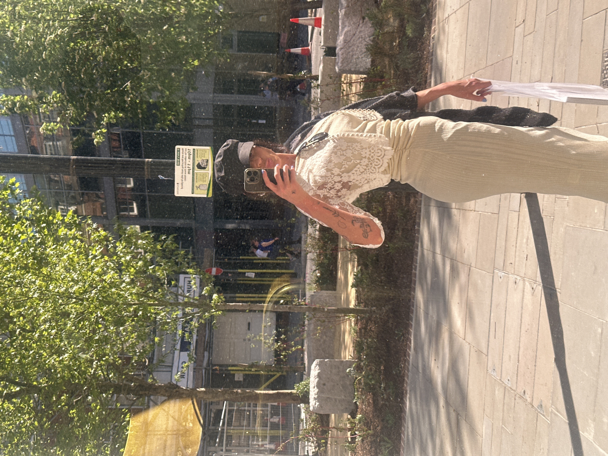 The image appears to be a reflection in a window or glass surface. A person is seen holding a smartphone, possibly taking a selfie. They are wearing a light-colored dress with lace detailing on the sleeves and a dark cap. They have visible tattoos on their arm. The reflection shows a cityscape with trees, buildings, and street furniture, suggesting an urban setting. There are traffic cones and construction materials visible in the background, hinting at nearby road work or construction. The scene has ample natural light. Based on the architectural style and presence of specific street furniture, it is likely to be a street in London, United Kingdom.