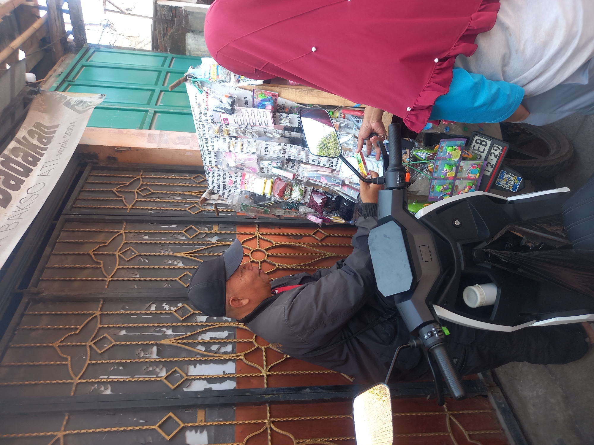 In Linggar, Indonesia, a man wearing a black baseball cap and a dark jacket is seen sitting on a scooter. He is looking to his right, seemingly engaged in a transaction with a person whose back is to the camera, wearing a red hijab. In front of them, a display rack is filled with various small items, possibly goods for sale, including what appear to be hair accessories and small toys. The background shows a building with a green door and an ornate metal gate. The scene appears to be an outdoor marketplace or street vendor setup.