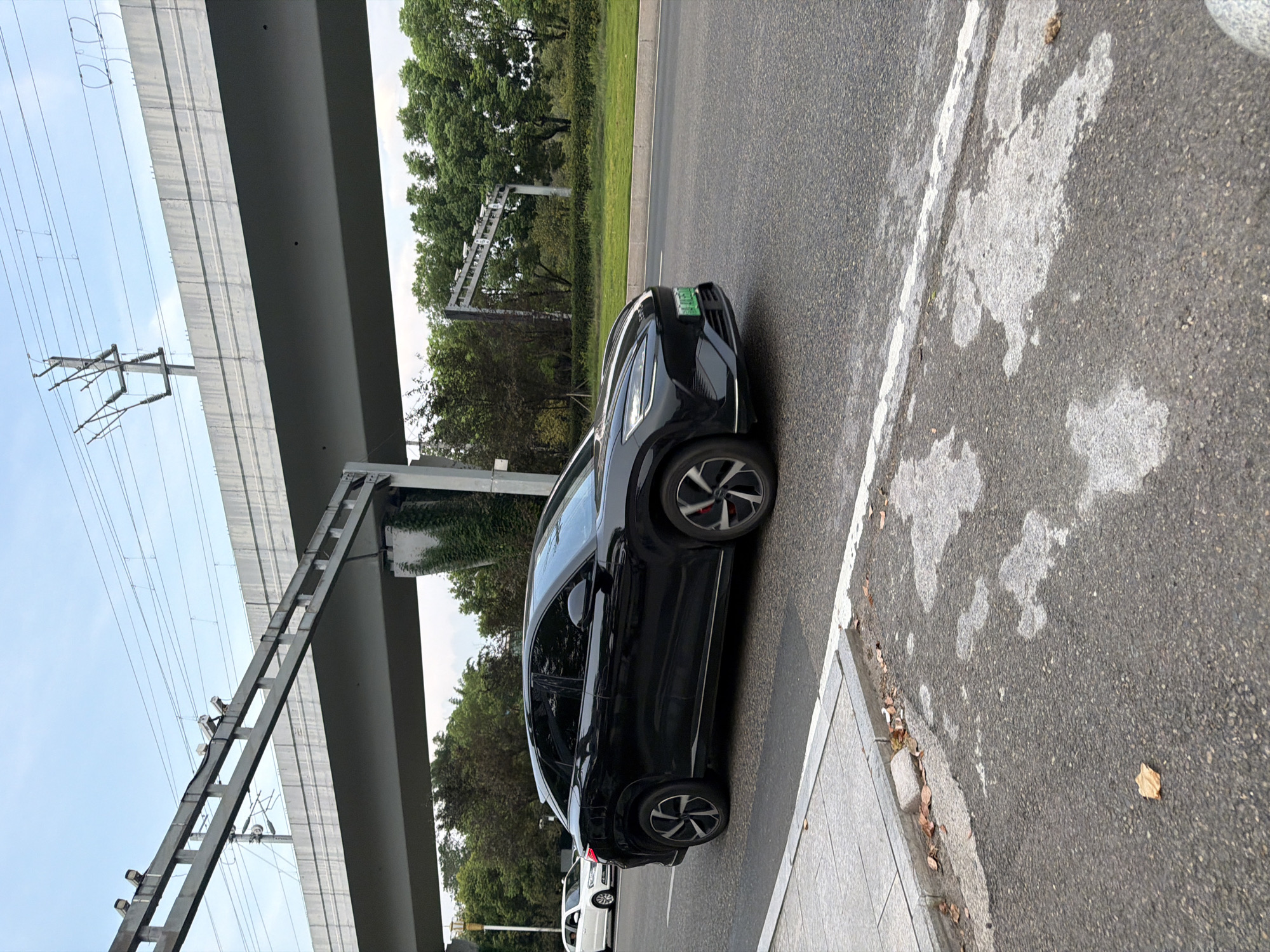 In Jiangjin, China, a black electric SUV with a green license plate drives on an asphalt road. The car is positioned in the foreground, angled towards the right of the frame. To the left of the road is a paved pedestrian walkway bordered by a concrete curb. Behind the walkway, a white sedan is partially visible. Elevated infrastructure, including what appears to be a highway overpass with overhead wires and traffic cameras, dominates the upper portion of the image. Lush green trees line the background beyond the road and the overpass. The sky is a pale blue with scattered clouds. The scene is captured during daylight hours.