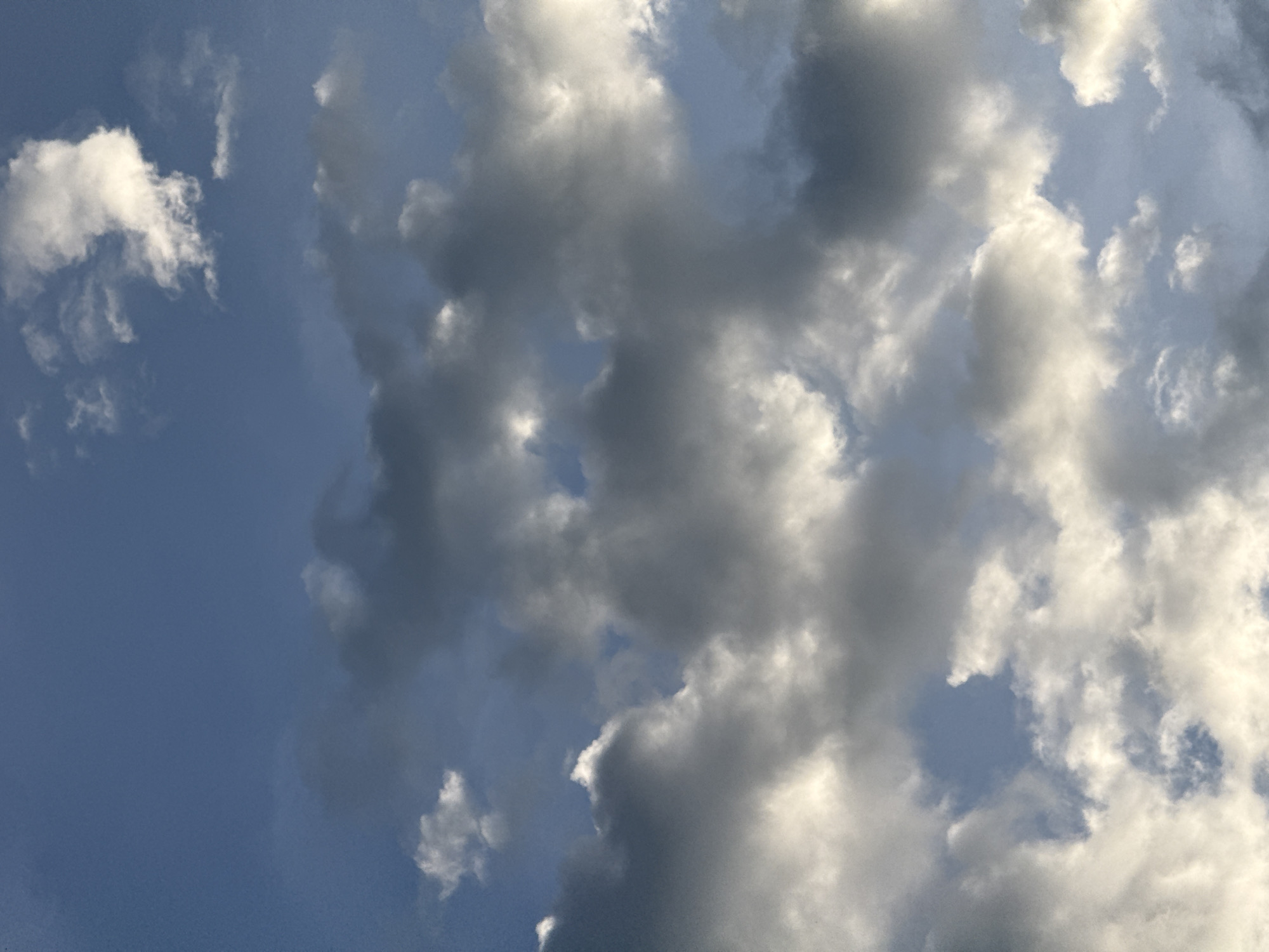 The image is a low-angle shot of a cloudy sky with patches of blue visible. The clouds are a mix of white and dark gray, suggesting a dynamic weather pattern. The lighting indicates it is either sunrise or sunset, with sunlight illuminating the edges of some clouds. The scene is devoid of any human presence, man-made structures, or terrestrial elements, depicting only the sky and clouds. The image does not contain any information that can definitively place it in Đà Nẵng, Vietnam.