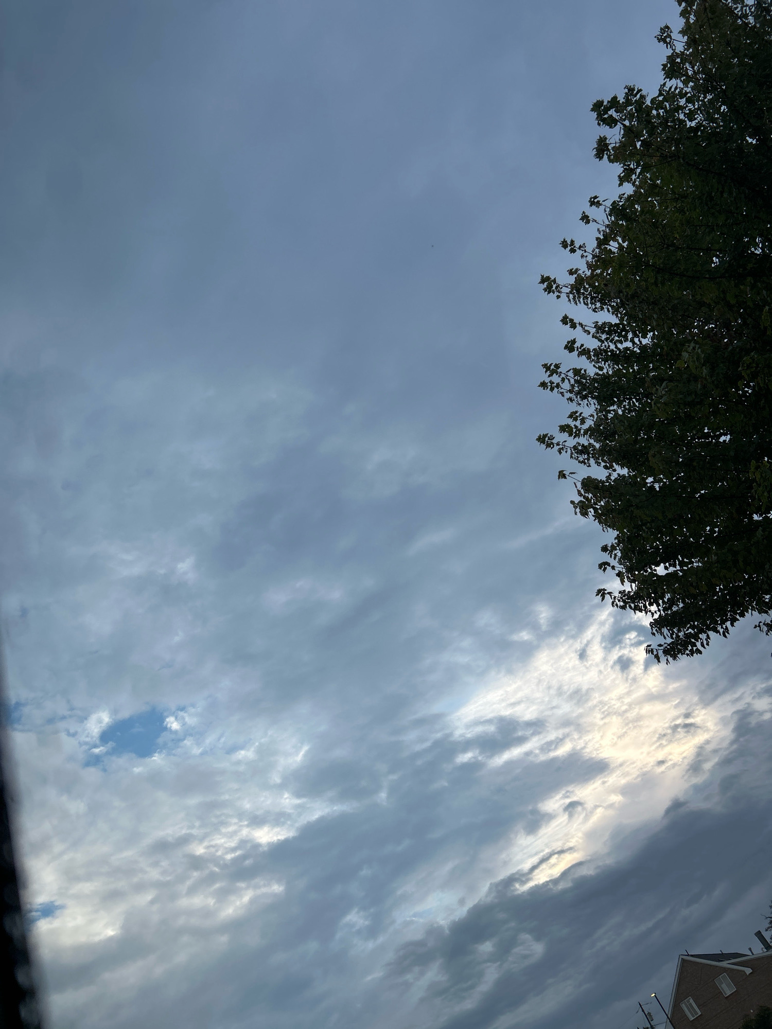 A portion of a tree with green leaves is visible in the upper right quadrant of the image. The sky is filled with gray and white clouds, with patches of blue sky peeking through. The sun is partially obscured by the clouds, casting a soft glow on the scene. In the lower right corner, the roof of a building with white-trimmed windows can be seen. The image appears to be taken outdoors in Robinson Township, United States of America.