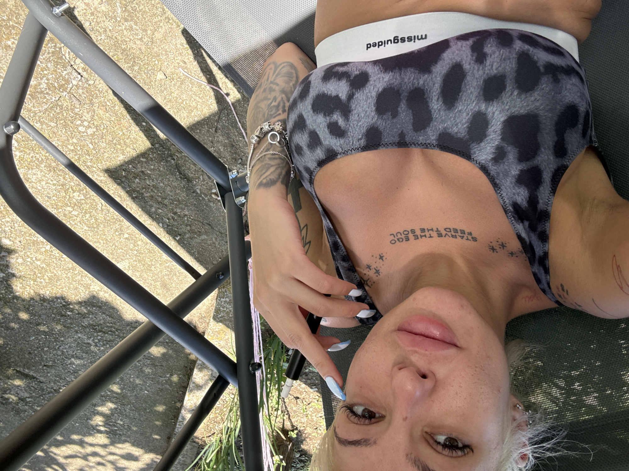 A blonde woman with short hair, fair skin, and light makeup is lying on a lounge chair outdoors. She is wearing a leopard-print sports bra with a white band that says "missguided." She has tattoos on her chest and arm. Her left hand, with light blue and white manicured nails, is holding an electronic cigarette. The background shows concrete, some greenery, and the frame of a lounge chair. The scene is lit by sunlight, casting shadows on the ground.