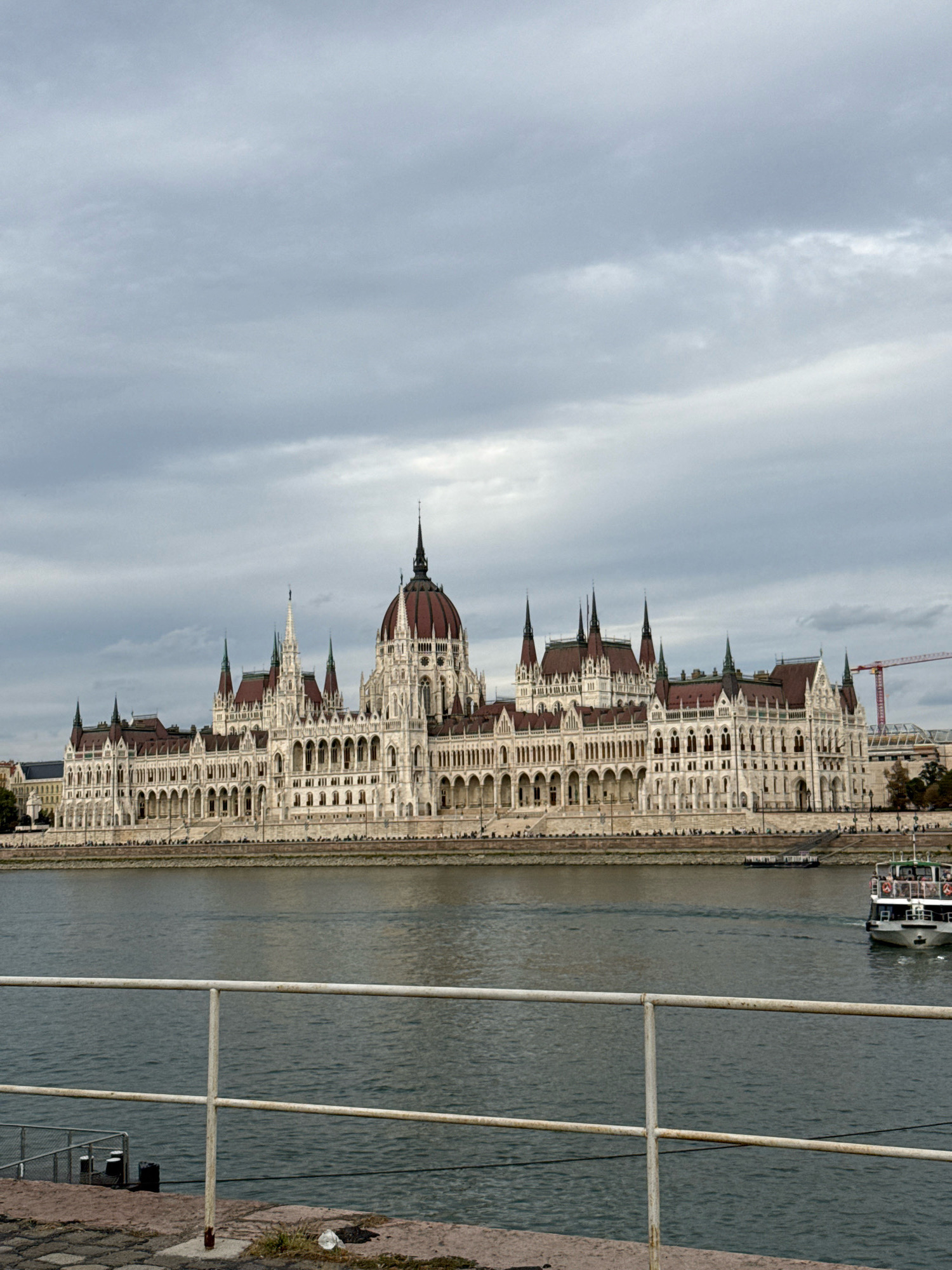 The image shows a wide shot of the Hungarian Parliament Building in Budapest, Hungary, viewed from across the Danube River. The building is grand and ornate, with a prominent dome and numerous spires, constructed from white stone with red roofs. The river in the foreground is a dark, choppy gray, with a white tourist boat in the middle distance heading away from the viewer. A white metal railing runs along the bottom of the frame, indicating a viewpoint from a promenade or walkway. The sky is overcast with heavy, gray clouds, suggesting a cool or rainy day. The ground in the foreground is paved with cobblestones and some reddish-brown stone.