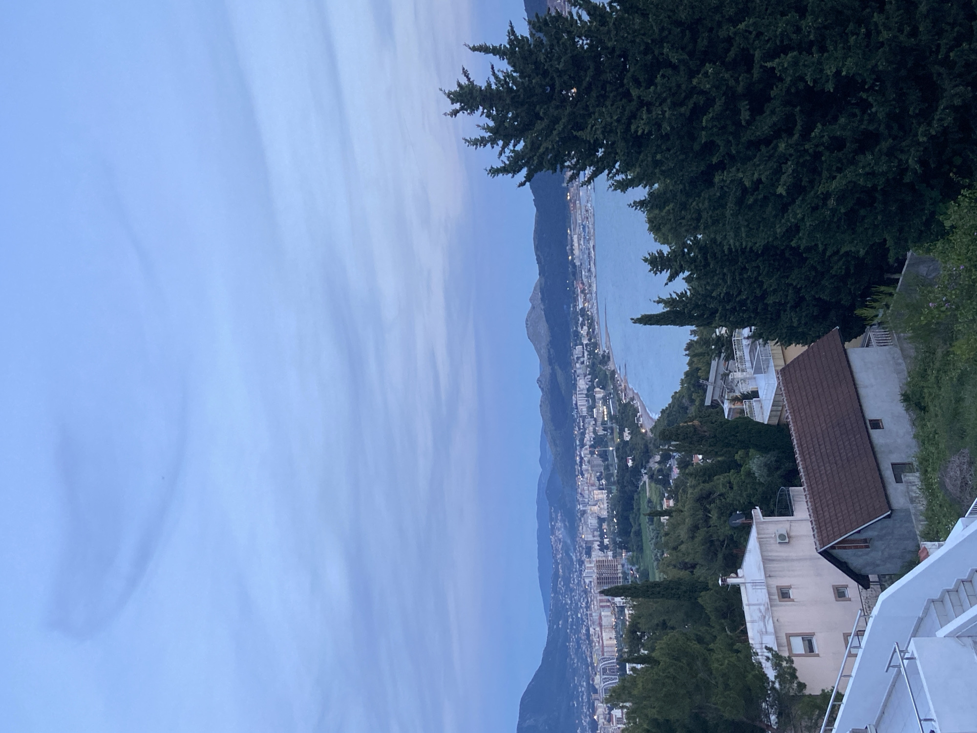 The image depicts a panoramic view of the city of Šušanj, Montenegro, situated along the Adriatic coastline. The photograph is taken from an elevated vantage point, possibly a hillside or a high-rise building, overlooking the cityscape and the sea. The sky appears to be in the early morning or late evening, indicated by the soft hues of blue and streaks of cloud formations.
In the foreground, residential buildings with red tile roofs and white walls are visible, suggesting a Mediterranean architectural style. Lush green trees and vegetation cover the slopes, adding a natural element to the urban environment. A set of white stairs leads downward, implying a path or access point from the viewpoint.
The city of Šušanj stretches along the coast, with densely packed buildings and infrastructure. The coastline curves gently, with a beach and harbor area noticeable. A mountain range rises in the background, providing a dramatic backdrop to the coastal city. The Adriatic Sea extends to the horizon, its surface reflecting the ambient light. The scene is quiet and tranquil, with no apparent human activity in the broader landscape.