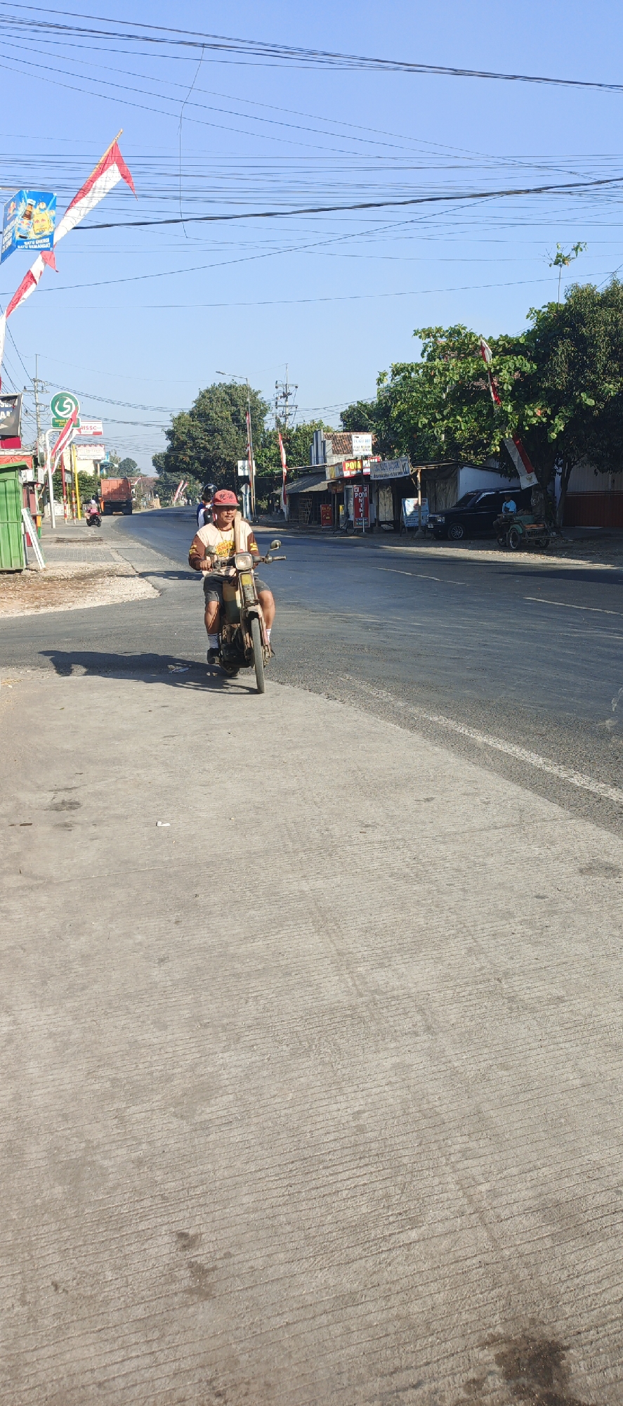 A man is riding a vintage motorcycle on the side of a road in Kasiyan, Indonesia. The man is wearing a red cap, a light-colored t-shirt with graphics, and shorts. He is looking forward and appears to be in motion. The motorcycle is a light brown color and appears to be an older model. The road is paved and appears to be a main thoroughfare with traffic and businesses on the right side. The environment is sunny with a clear blue sky. There are trees and buildings visible along the road, and Indonesian flags are flying from poles. The pavement in the foreground shows tire marks and some minor debris.