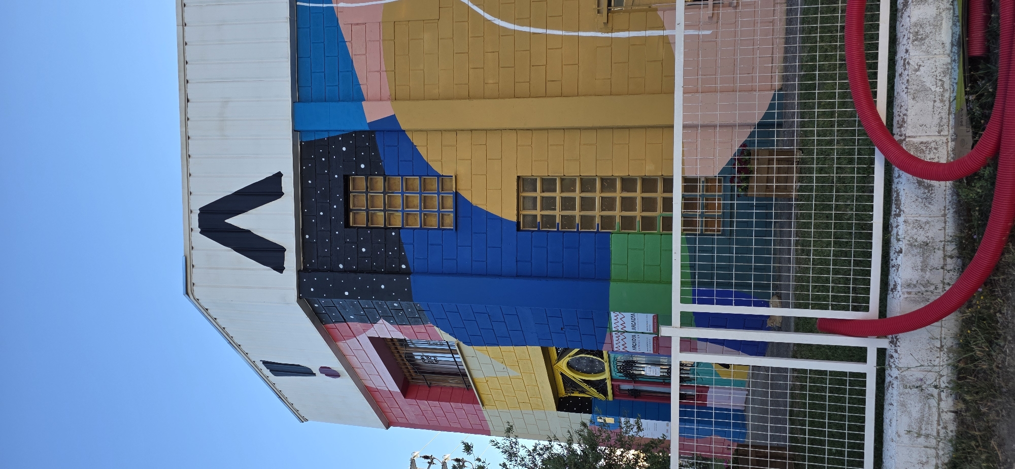 The image shows a brightly painted building in Huesca, Spain, with abstract geometric shapes and patterns in vibrant colors like blue, yellow, pink, and red. A large black "A" shape is painted on the white upper section of the building, near the roofline, with a black exclamation mark also visible on the side. The building has several windows, some of which are made of textured glass blocks. A white metal fence with square mesh is in the foreground, partially obscuring the lower part of the building. Red corrugated pipes are lying on the ground in front of the fence. The sky above is clear and blue.