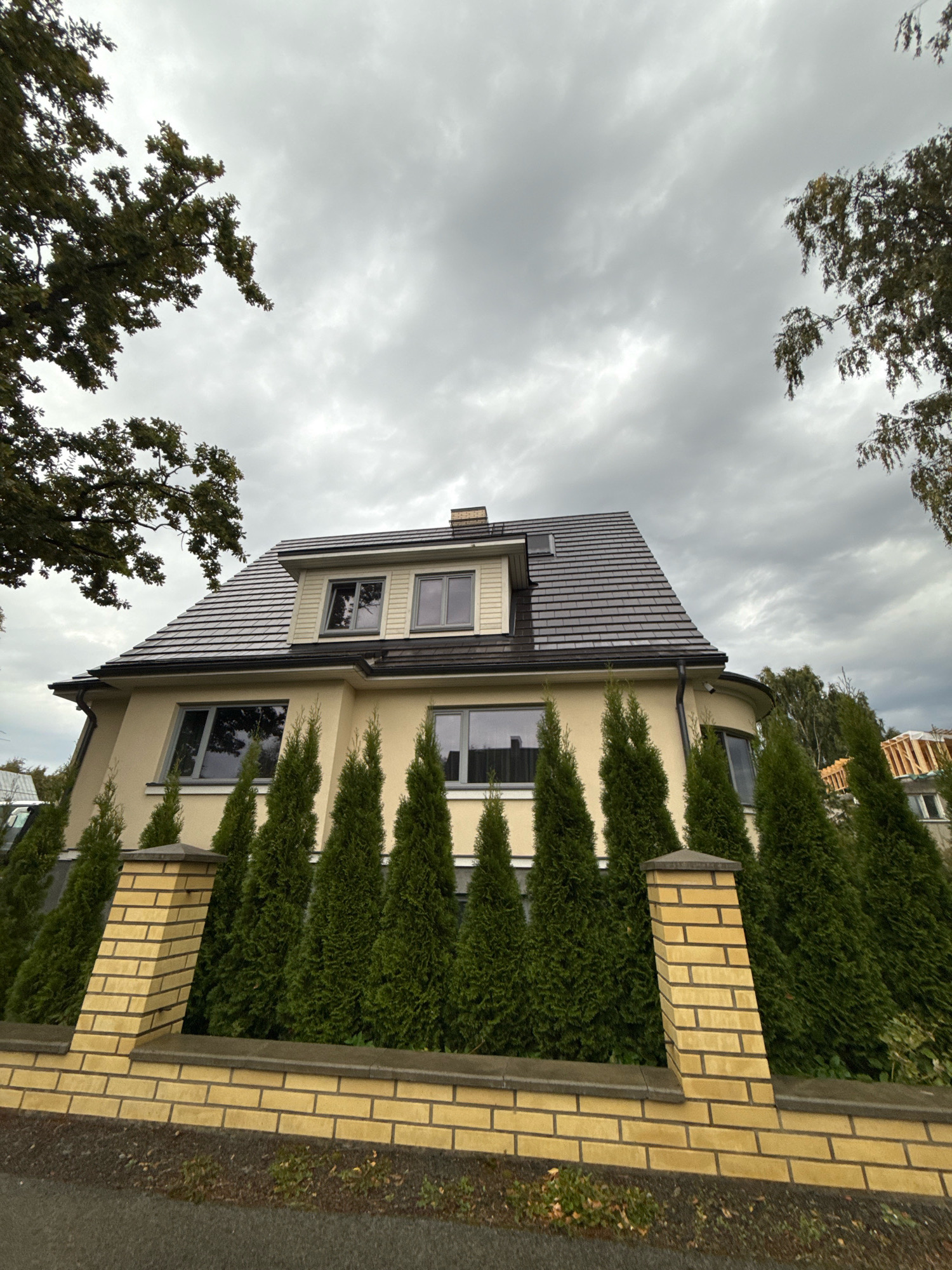 A low-angle, wide shot captures a modern, two-story house with a light yellow facade and a dark gray tiled roof. The house features several large, rectangular windows with dark frames. A row of tall, conical evergreen trees stands in front of the house, partially obscured by a yellow brick fence with dark gray capping. The fence has decorative angled pillars. The sky above is overcast with heavy, gray clouds. In the background, more trees are visible, and on the right side, the upper stories of another building under construction can be seen. The foreground shows a patch of dark asphalt and some sparse green ground cover. The image appears to have been taken outdoors in a suburban or residential area of Tallinn, Estonia.