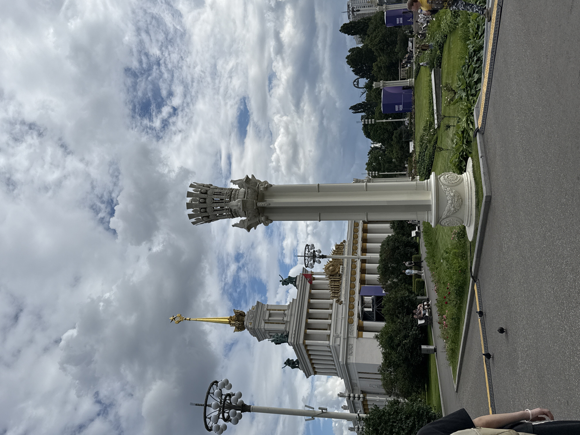 This media file captures a sunny day at an outdoor exhibition, likely in Moscow, Russia, given the architectural style. The main subjects are a grand, white neoclassical building with a golden spire, a tall decorative column, and lush green gardens. Several people are visible in the background, strolling through the gardens and sitting on benches. There are also decorative lamps and sculptures on the building. The sky is a mix of white clouds and blue sky, indicating fair weather. The overall mood is peaceful and relaxed, typical of a day at a public exhibition or park.