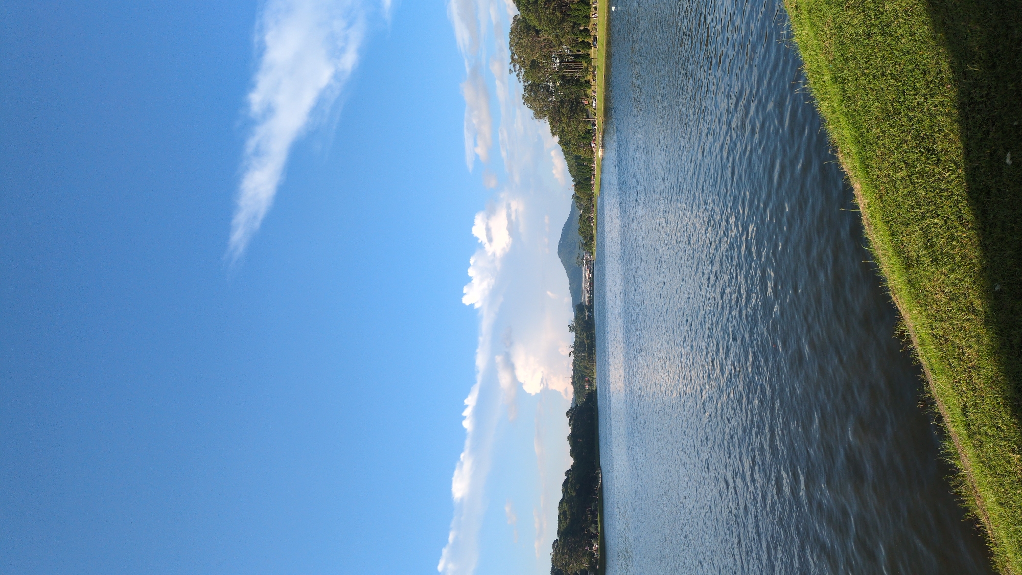 This image depicts a serene view of Xuan Huong Lake in Da Lat, Vietnam, under a bright blue sky dotted with scattered white clouds. The foreground features a lush green grassy embankment sloping down to the dark, rippling water of the lake. Across the water, the middle ground is occupied by a treeline with dense green foliage, and further in the distance, a prominent, cone-shaped mountain rises against the horizon. A faint bridge or structure can be observed spanning across the water in the far distance. The overall scene conveys a peaceful, natural landscape within the city.
