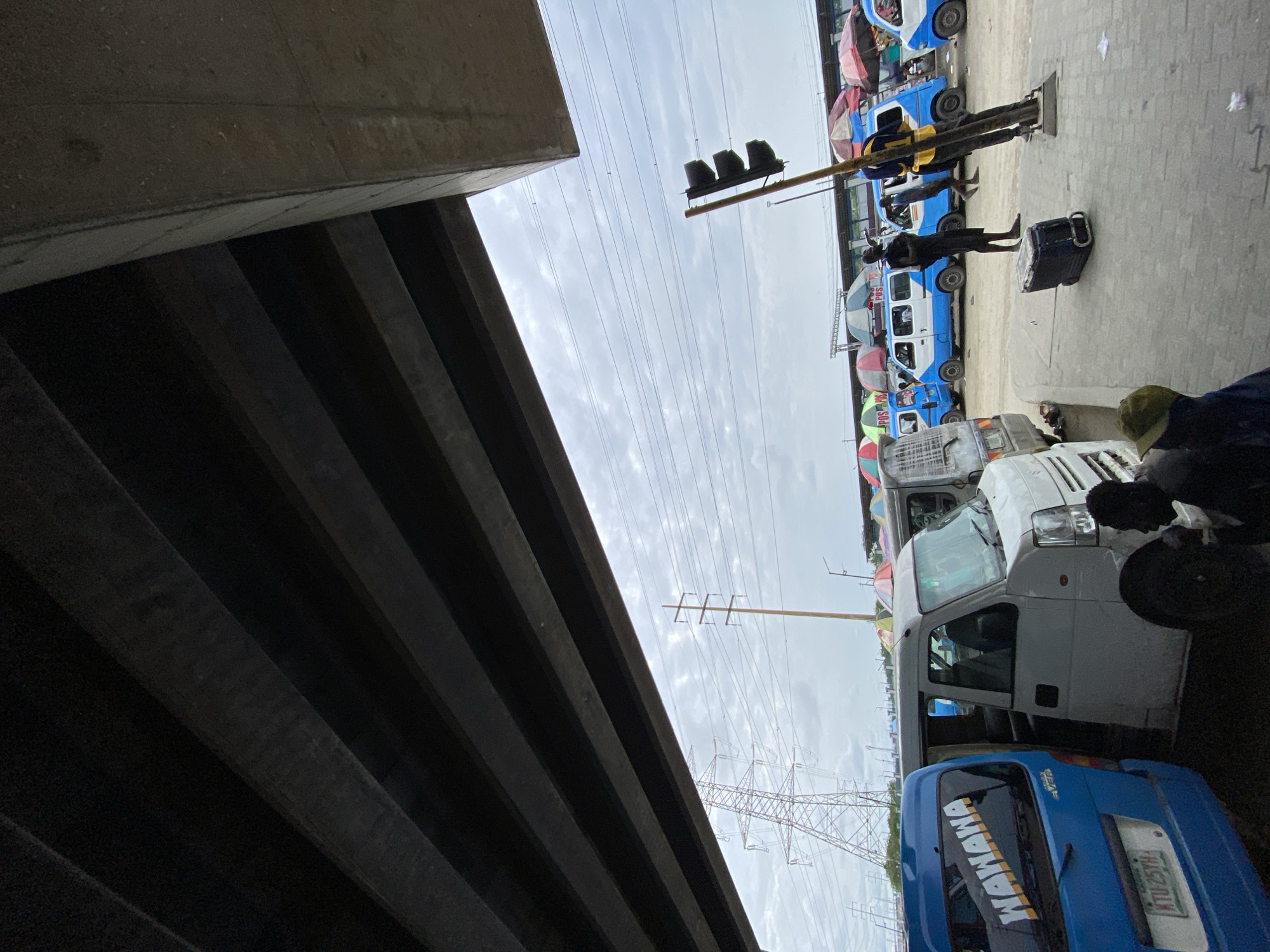 The image shows a street scene in Lekki, Nigeria. The view is from under a concrete overpass looking toward a row of parked mini-buses. Some of the buses are blue and white, others are gray. There is a traffic light suspended above the vehicles. People are standing beside the buses. An electrical tower is visible in the background.