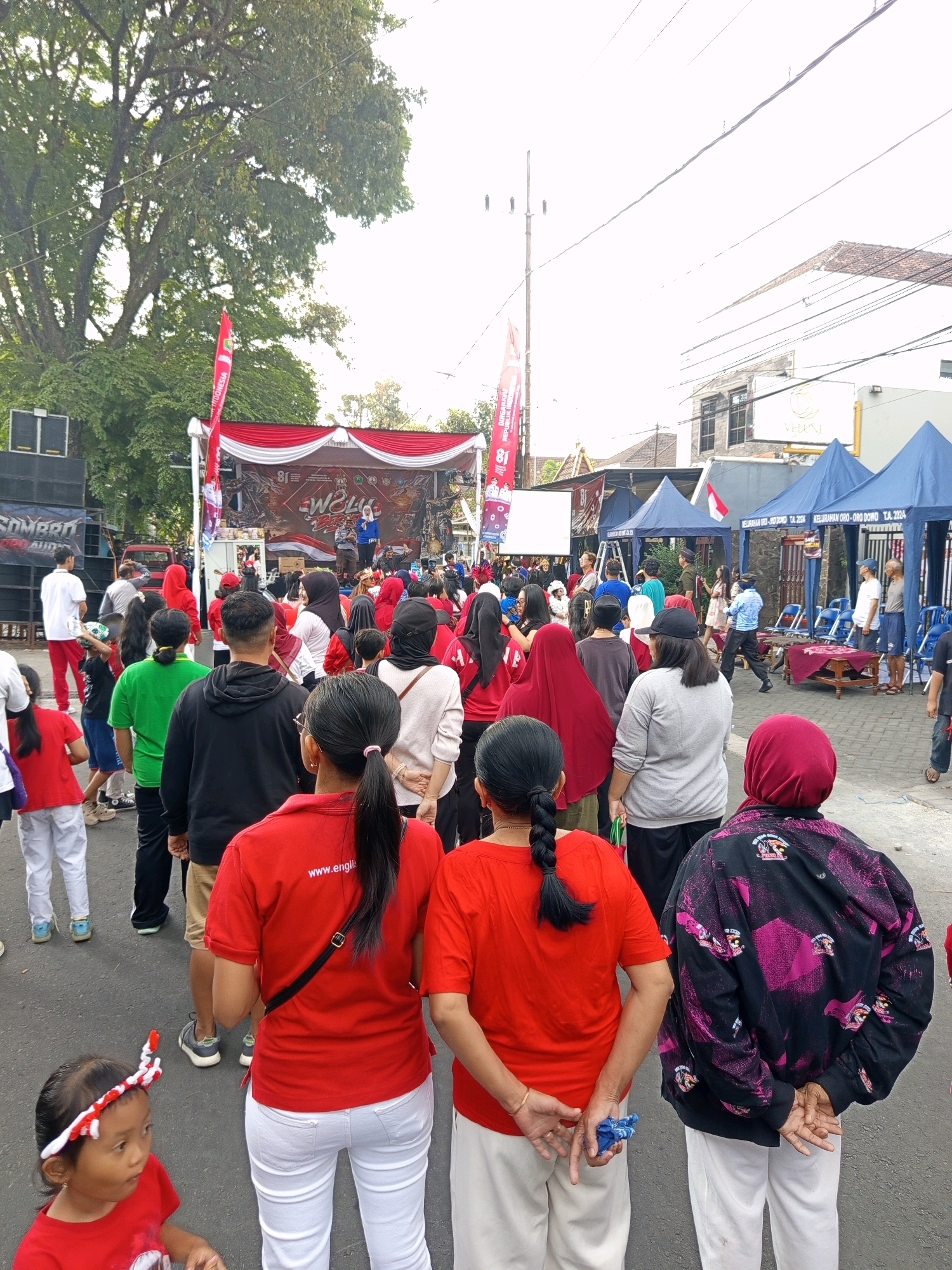 The image depicts a street scene in Malang, Indonesia, with a crowd gathered for an event. In the background, a stage is set up with a red and white banner, and several people are on stage, one of whom is speaking into a microphone. To the left of the stage, a large speaker system is visible. The street is lined with tents on the right side, and some people are seated in chairs in front of them. The crowd consists of many individuals, mostly dressed in red and white attire, suggesting a patriotic or celebratory theme. Some people are standing and watching the stage, while others are walking. A young girl in the foreground is wearing a red outfit and a red and white headband. The overall atmosphere appears to be lively and communal.