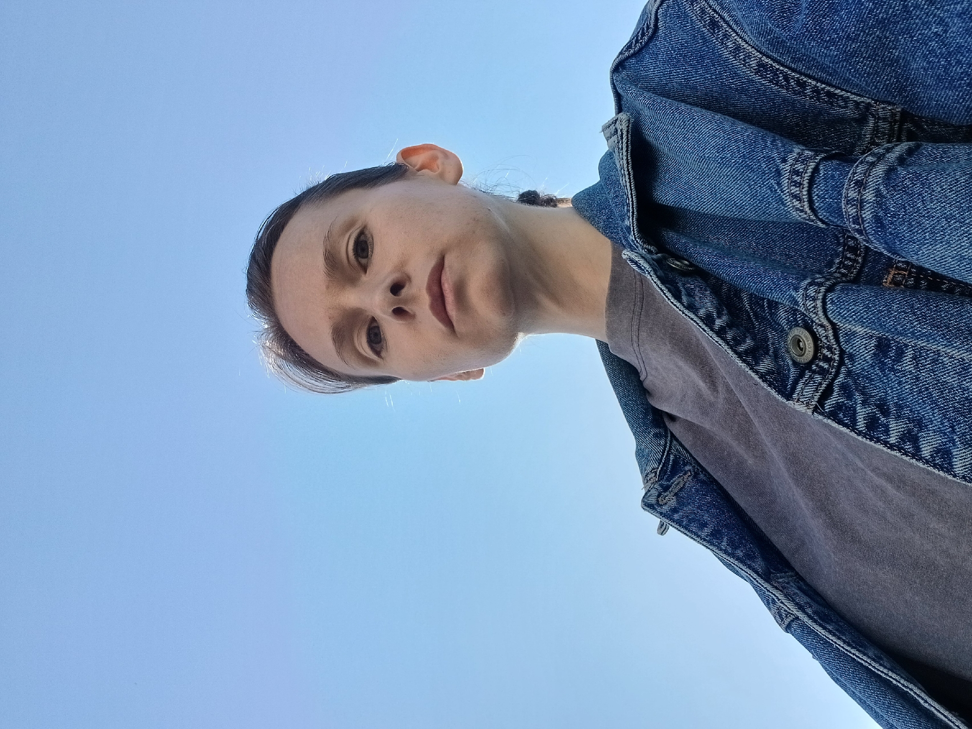 This is a selfie taken from a low angle looking up at the sky, with a person's face and upper body visible in the frame. The person appears to be looking directly at the camera with a neutral expression. They are wearing a denim jacket over a grey t-shirt. The background is a clear blue sky, suggesting it is daytime and likely good weather. The image has a slightly tilted orientation. No visible text is present. The time of day appears to be daytime given the bright sky. The setting is outdoors, indicated by the clear sky. The location context provided is Yekaterinburg, Russia, which is not directly discernible from the image itself. The overall mood is straightforward and unpretentious.