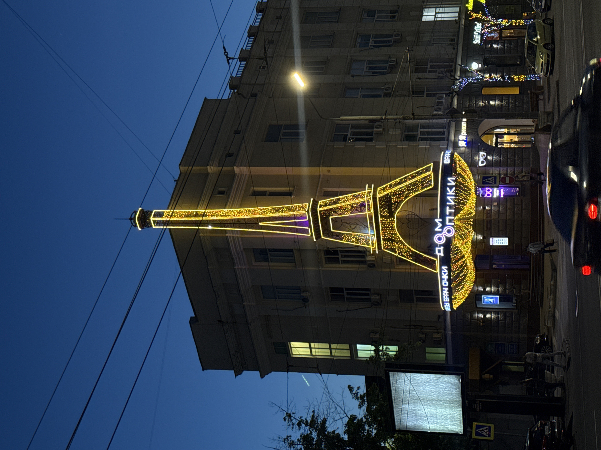 The image shows a nighttime street scene in Rostov-on-Don, Russia. A brightly lit Eiffel Tower replica stands in front of a building with lit windows. The building houses "ДОМ ОПТИКИ" (Optics House), indicated by vertical signage. The sky is a deep blue, transitioning to a lighter blue near the horizon. Power lines crisscross the upper part of the image. A car with illuminated rear lights is visible on the right side of the frame, suggesting traffic on the road. A pedestrian crossing sign is visible, and there appears to be a tree in the foreground. Additional street lights and building lights illuminate the scene.