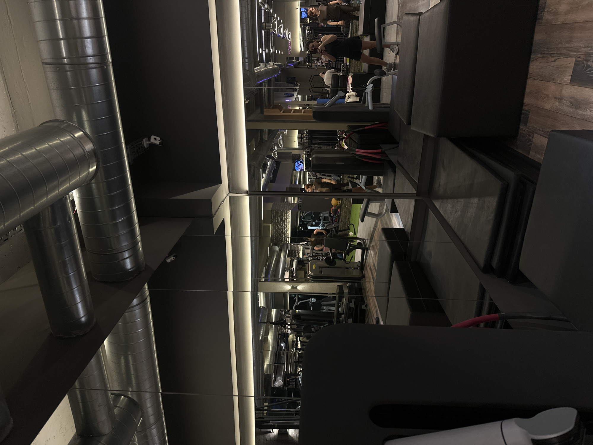 The image shows the interior of a gym with mirrored walls, creating a long, repetitive visual effect. The gym equipment is visible in the reflection, including weight machines and workout benches. Several people are present, some using the equipment and others standing or walking. Industrial-style metal ventilation ducts run along the ceiling. The location is identified as a gym in Yerevan, Armenia.