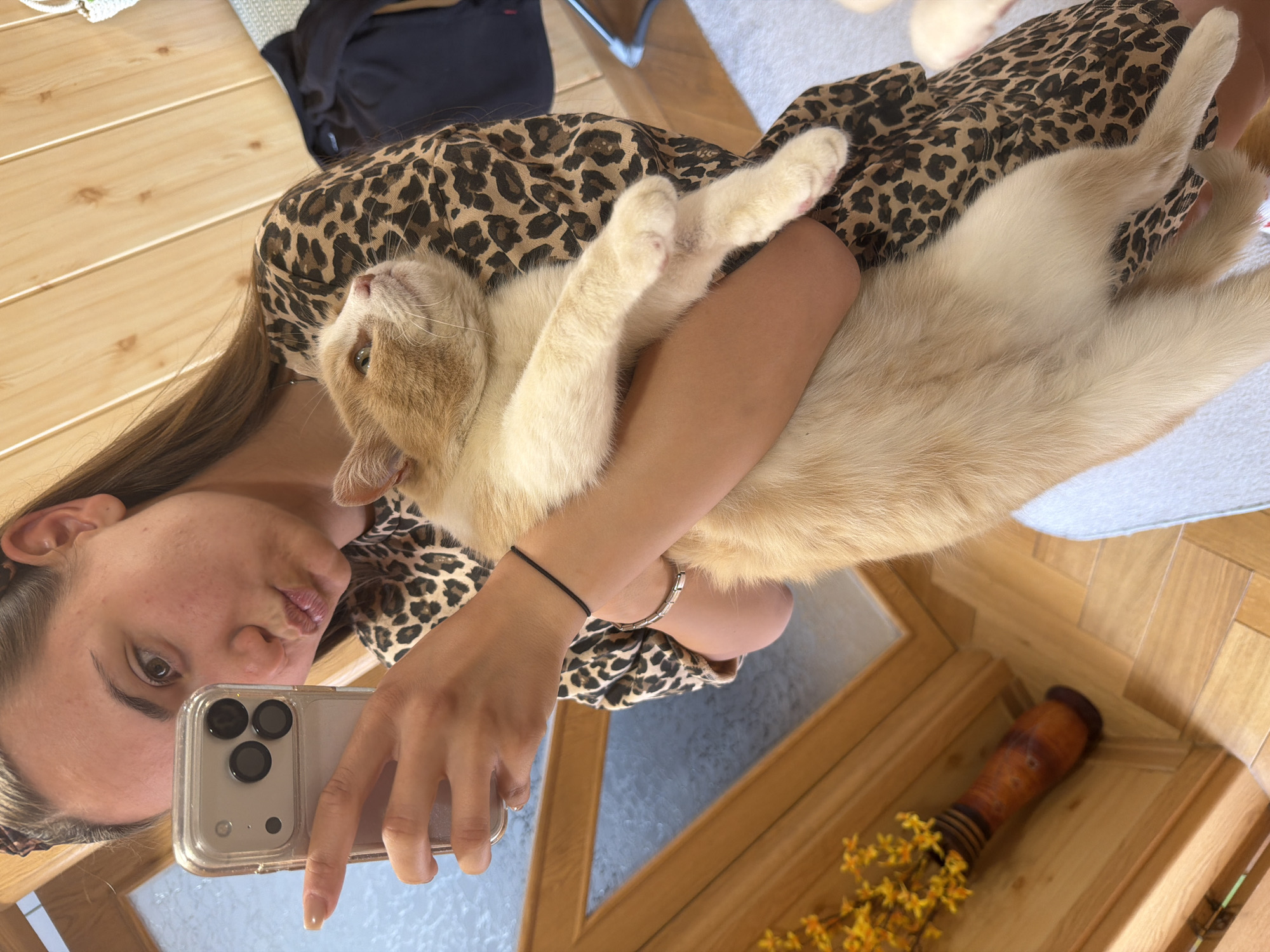 A woman in a leopard print dress is holding a light orange cat, possibly indoors. The woman is holding up a phone to take a selfie. Her hair is long and brown. She is wearing a black bracelet on her left wrist and a silver bracelet on her right wrist. The cat is in her arms with its legs extended. The location is likely not in Buhovo, Bulgaria, as the setting appears to be indoors in a home, which doesn't allow for geolocation. There is a wooden floor, a table with a wooden frame, and a vase with yellow flowers near the table's edge. A rug or other covering is partially visible in the background.