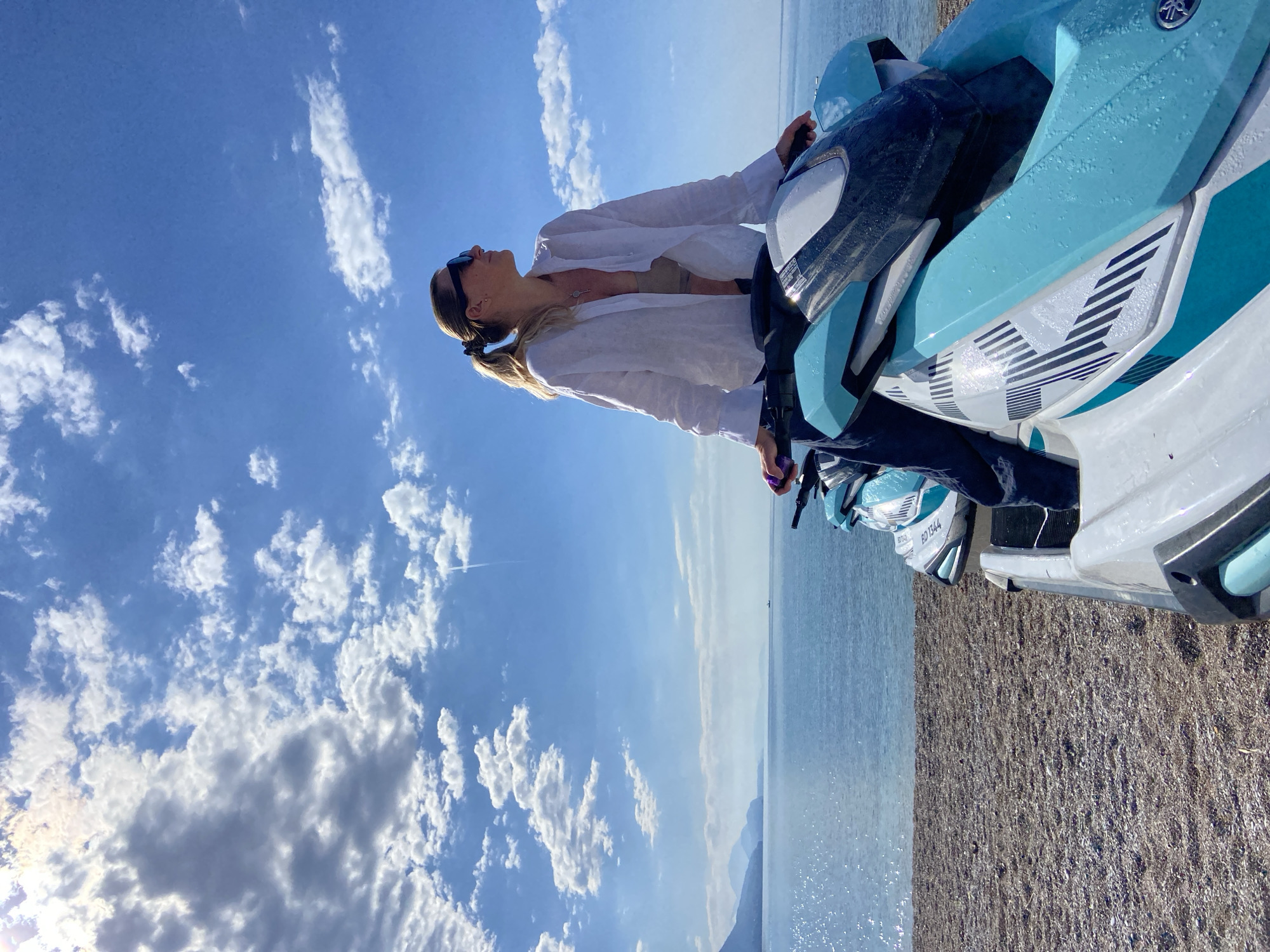 A vertical shot reveals a sunlit outdoor scene featuring a woman atop a blue and white Yamaha jet ski. The location is on a beach in Prijevor, Montenegro, as evidenced by the visible boat registration number (BD 1344) on an adjacent jet ski. The woman, wearing sunglasses and a white linen shirt with a light-colored top underneath, looks upwards. The background showcases a sky with cumulus clouds and the tranquil Adriatic Sea. The foreground displays a stretch of shoreline with fine gravel.