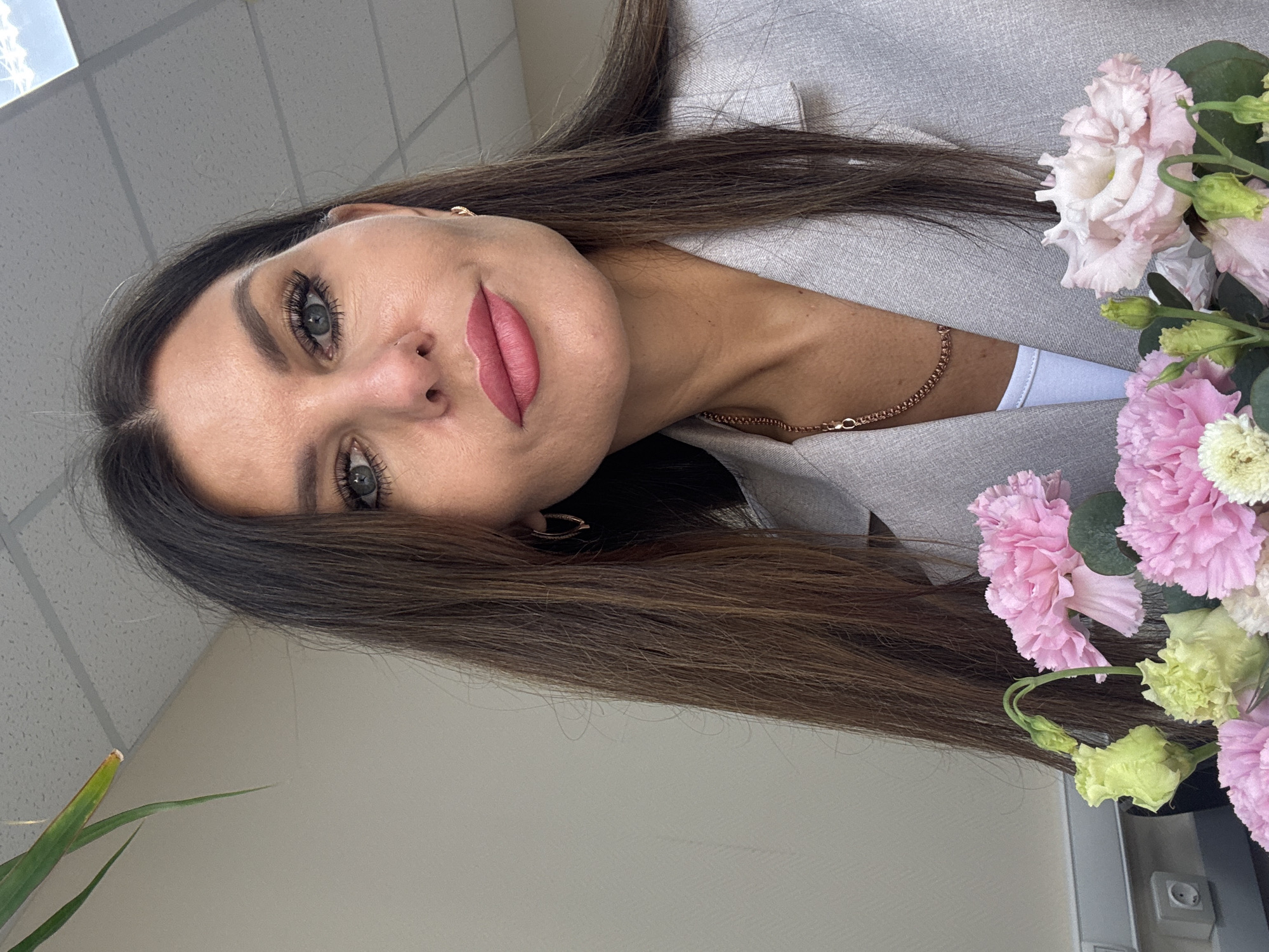 A medium close-up shot captures a woman with long, dark brown hair and striking blue-grey eyes. She has a fair complexion and wears pink lipstick. She is dressed in a grey blazer, with a hint of a white top visible underneath. She has gold earrings and a delicate gold chain necklace. To her right, a bouquet of pink, white, and green flowers adds a vibrant touch. The background appears to be an interior space with a white ceiling with a square grid pattern and plain wall, possibly located in the city of Chaikovsky, Russia.