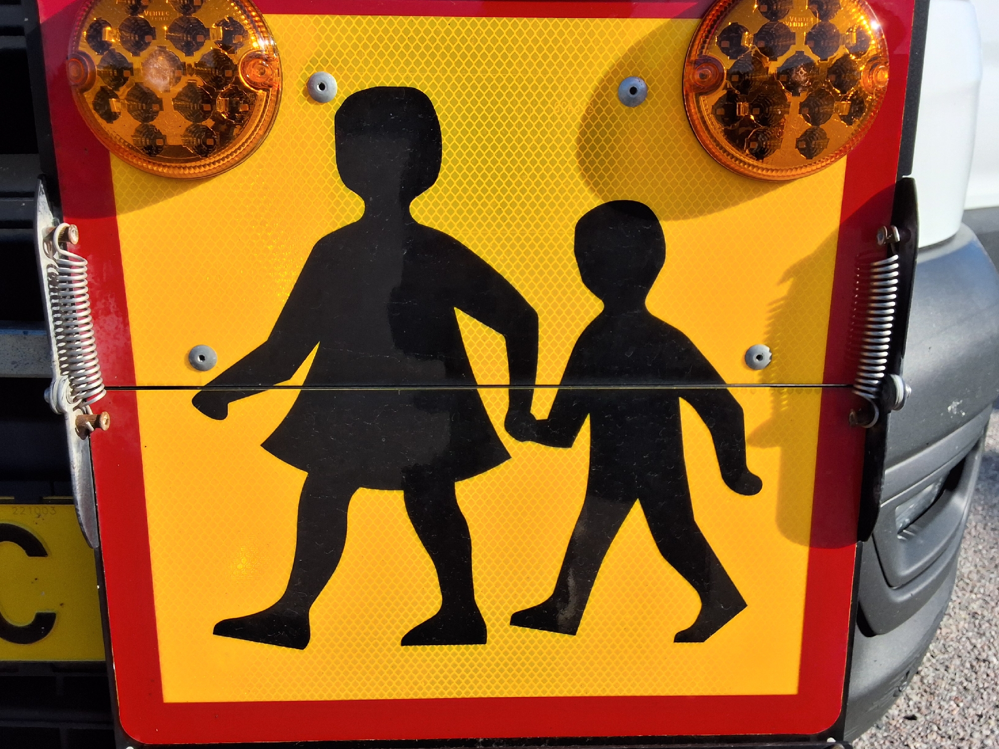 A close-up, eye-level shot captures a yellow school bus sign featuring two black silhouettes of children walking, one adult-sized and one smaller child-sized. The sign is framed by a red border. Two round, amber lights are mounted at the top of the sign, one on each side. Metal springs and fasteners are visible on the left and right edges of the sign, suggesting it is attached to a vehicle. A portion of the vehicle's grey bumper and a yellow license plate with the letter "C" are visible on the lower left. The sign is reflective, with a diamond pattern visible on its surface. The overall scene depicts a safety sign on a school bus in Arboga, Sweden.