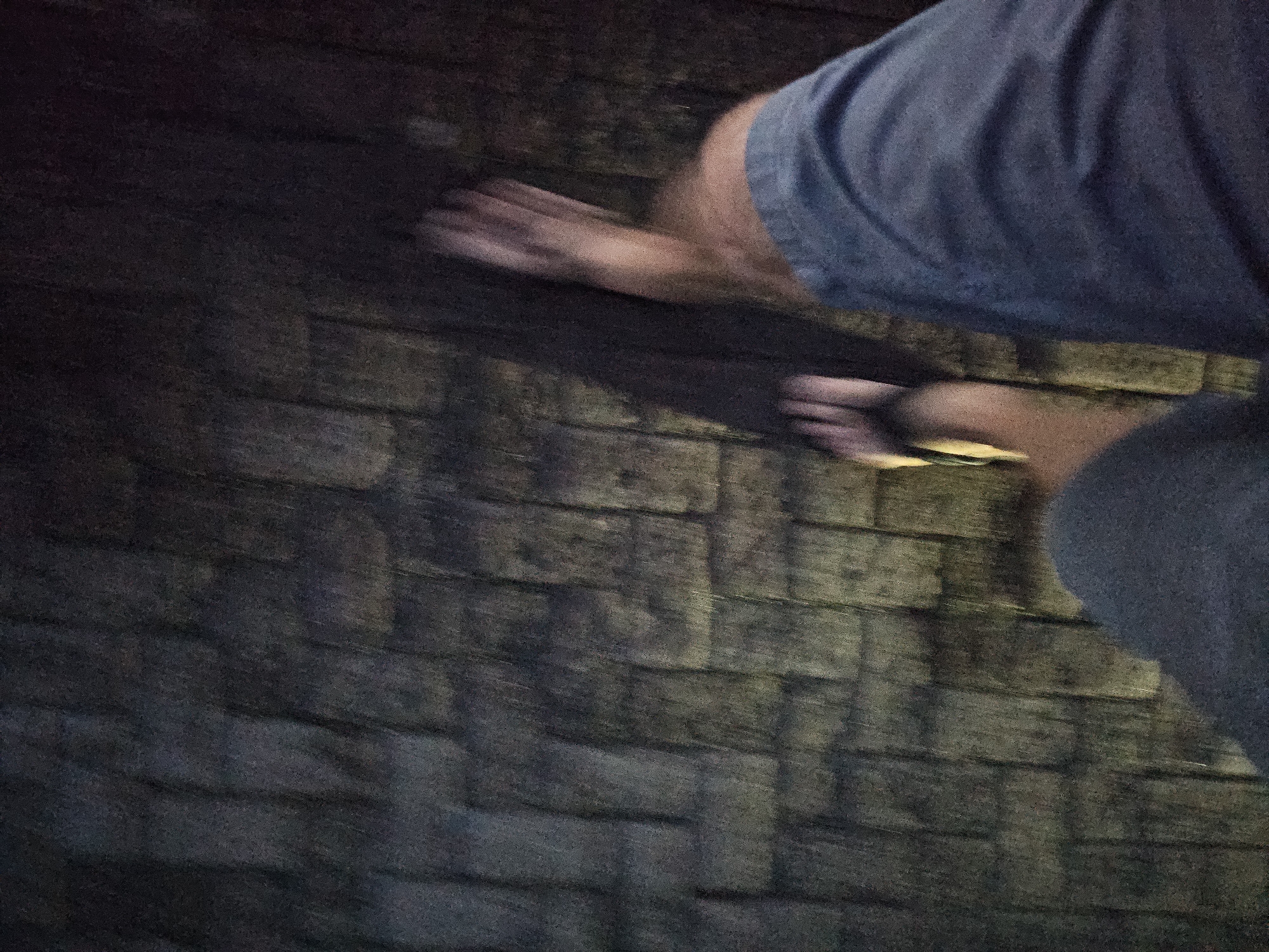 A person wearing a blue shirt is standing on a dark brick or stone surface. Their hand is blurred, suggesting movement. The scene appears to be dimly lit. The location is unknown.
