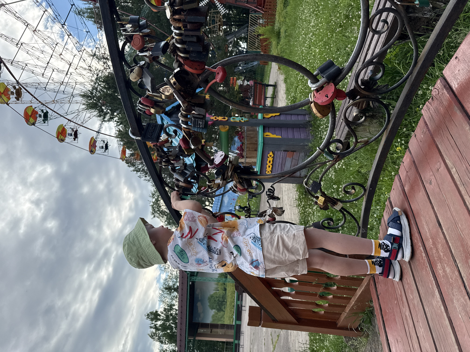 A young child wearing a light green hat, a white t-shirt with colorful graphics, and beige shorts stands on a wooden platform. The child is looking up at a metal structure adorned with numerous padlocks, some of which are heart-shaped. The child's feet are clad in blue and red sandals with white socks featuring yellow stripes and a black skeleton design. In the background, a Ferris wheel with red and yellow gondolas is visible, along with trees and a grassy area dotted with small white flowers. Behind the Ferris wheel, there are buildings with colorful decorations. The sky is partly cloudy, suggesting it's daytime. The scene appears to be an amusement park or a public recreational area. A notable detail is the presence of many padlocks, often symbolizing love or commitment. The overall mood seems peaceful and observational, with the child engrossed in examining the padlocks. There is some visible text on the t-shirt and on some of the padlocks. The text on the t-shirt includes phrases like "NEW," "Like," and "Dream," and "ACTIVE BOY." One padlock clearly shows "APECS." The location is contextually Chaikovsky, Russia.
