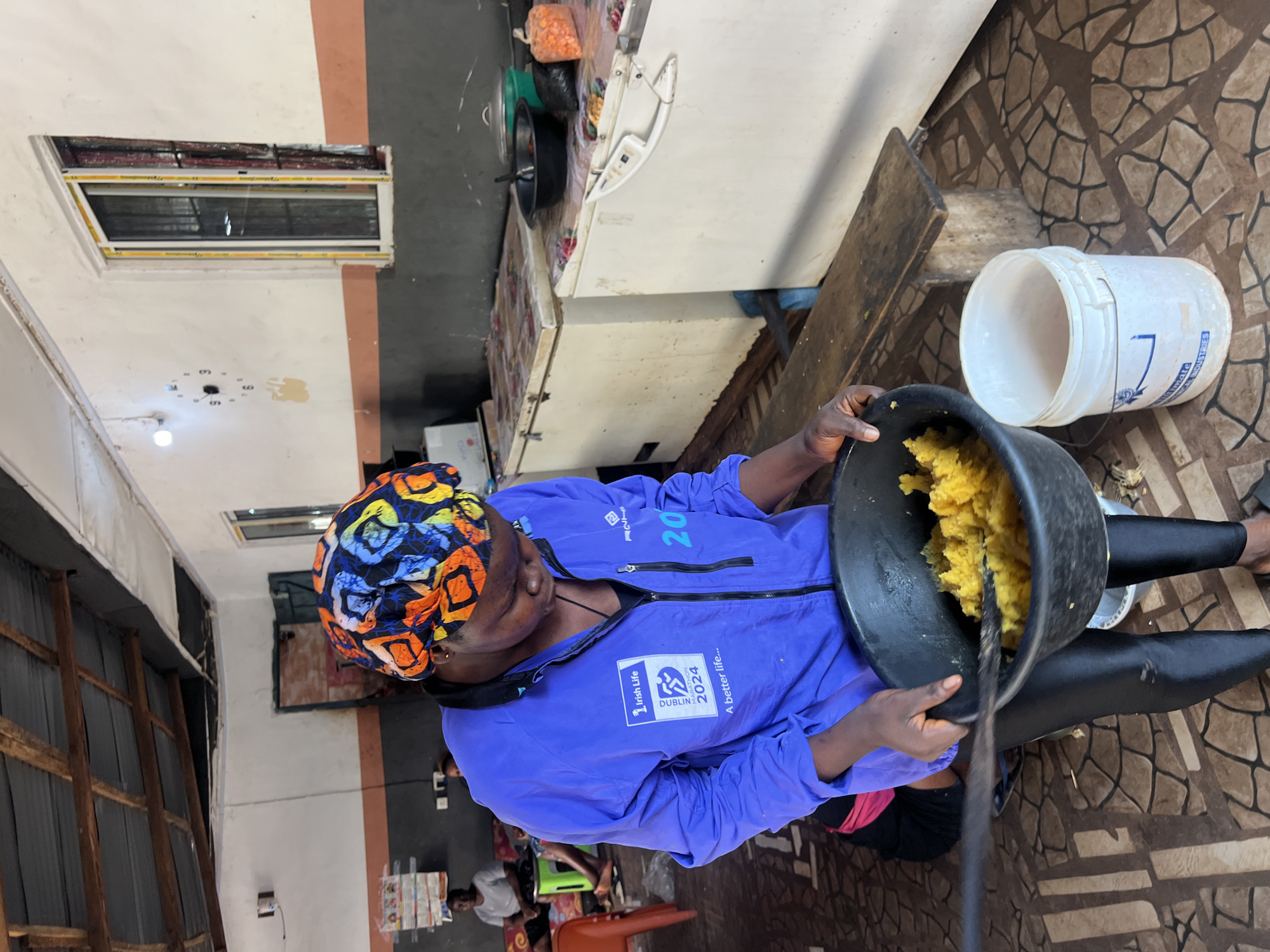 In Nkpor, Nigeria, a woman wearing a vibrant headwrap and a blue jacket with Irish Life and Dublin Marathon 2024 logos is stirring a large black bowl filled with yellow mashed food, possibly yams, using a dark utensil. She is standing on a tiled floor and in front of a white refrigerator. Behind her, other women are seated and the room appears to be a communal kitchen or dining area, with corrugated iron roofing and a clock on the wall. The environment suggests a modest, possibly institutional, setting.