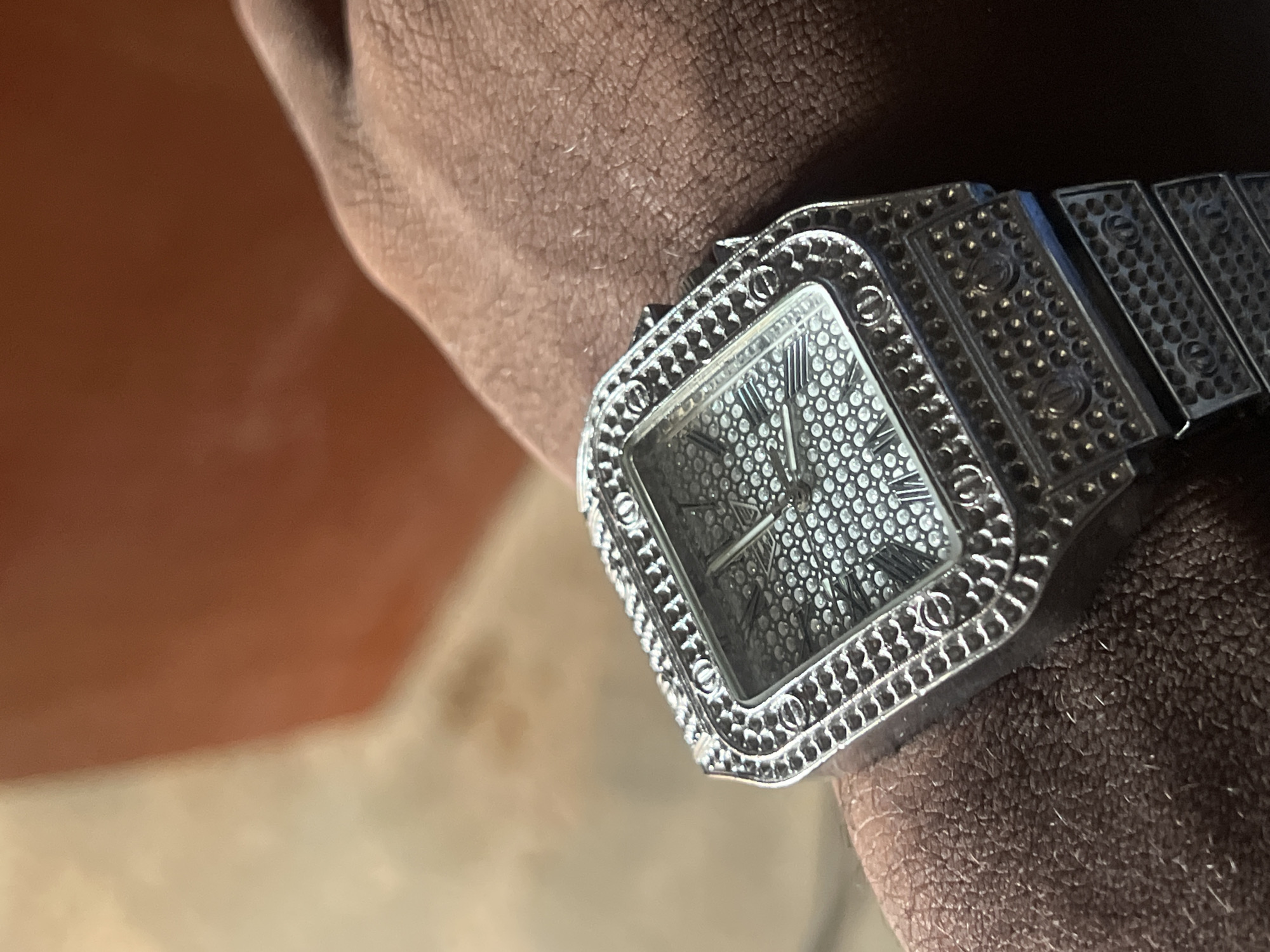 An iced-out, silver-toned, rectangular watch with Roman numerals on its face is worn on a person's wrist. The watch face and bezel are covered in small, circular, sparkling stones. The watch band is also made of silver-toned metal and features a textured pattern. The background is out of focus and appears to be an orange or brown surface. There is no discernible information about the location in the city of Wumat, Nigeria.