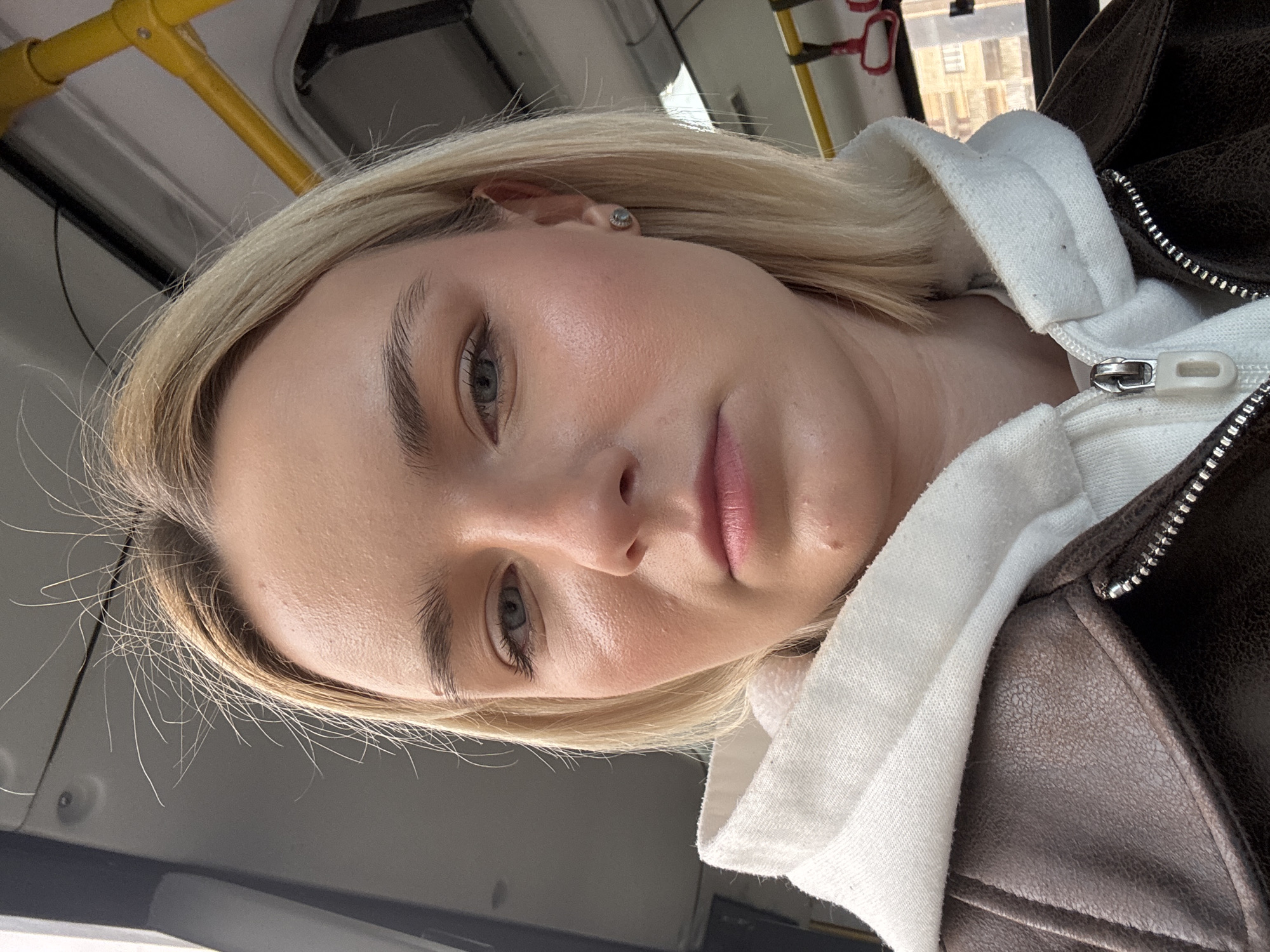A young woman with fair skin, blonde hair, and light-colored eyes is taking a selfie indoors. She is wearing a white hoodie with a brown jacket layered over it. The location appears to be inside a public transportation vehicle, possibly a bus or trolley. Outside the window, a building can be seen. It is in Vsevolozhsk, Russia.
