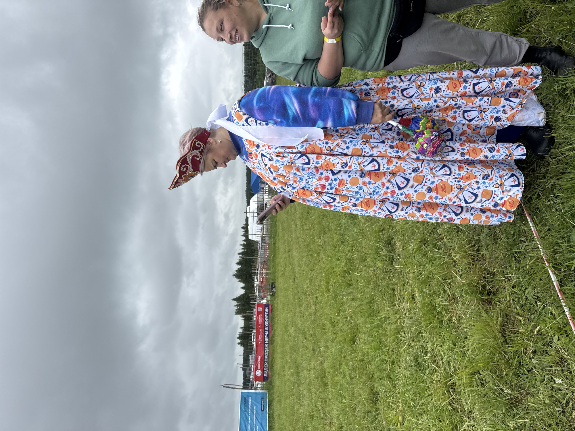 The image depicts two women standing in a grassy field under an overcast sky in Большая Кивара, Russia.
The woman on the left is an adult female with short, light pink hair, wearing a red kokoshnik-style headpiece with gold embroidery and a white sash. She is dressed in a long, multi-colored dress featuring a pattern of orange samovars and blue/red dishes over a blue and purple galaxy-print long-sleeved shirt. She holds a pink smartphone in her hands and looks down at it. In her right hand, she also holds a colorful, multi-lobed fabric toy or decoration. Her footwear consists of black shoes.
The woman on the right, also an adult female, has light brown hair pulled back. She wears a green hooded sweatshirt, grey sweatpants, and a black fanny pack around her waist. A yellow wristband is visible on her left wrist. She is looking to her left with a slight smile. Her footwear consists of black shoes and socks.
In the background, a green field extends with a line of evergreen trees in the distance. Several banners are visible on a wooden fence structure: a blue banner to the left and a prominent red banner that reads "НИКИТАН ЛИДЕР ПРОДАЖ МЕРЧА В УДМУРТИИ" (NIKITAN LEADER IN MERCHANDISE SALES IN UDMURTIA). Further back, white and blue tent-like structures are partially visible. The sky is uniformly grey and cloudy. A red and white striped pole or tape lies on the ground in the lower left foreground.