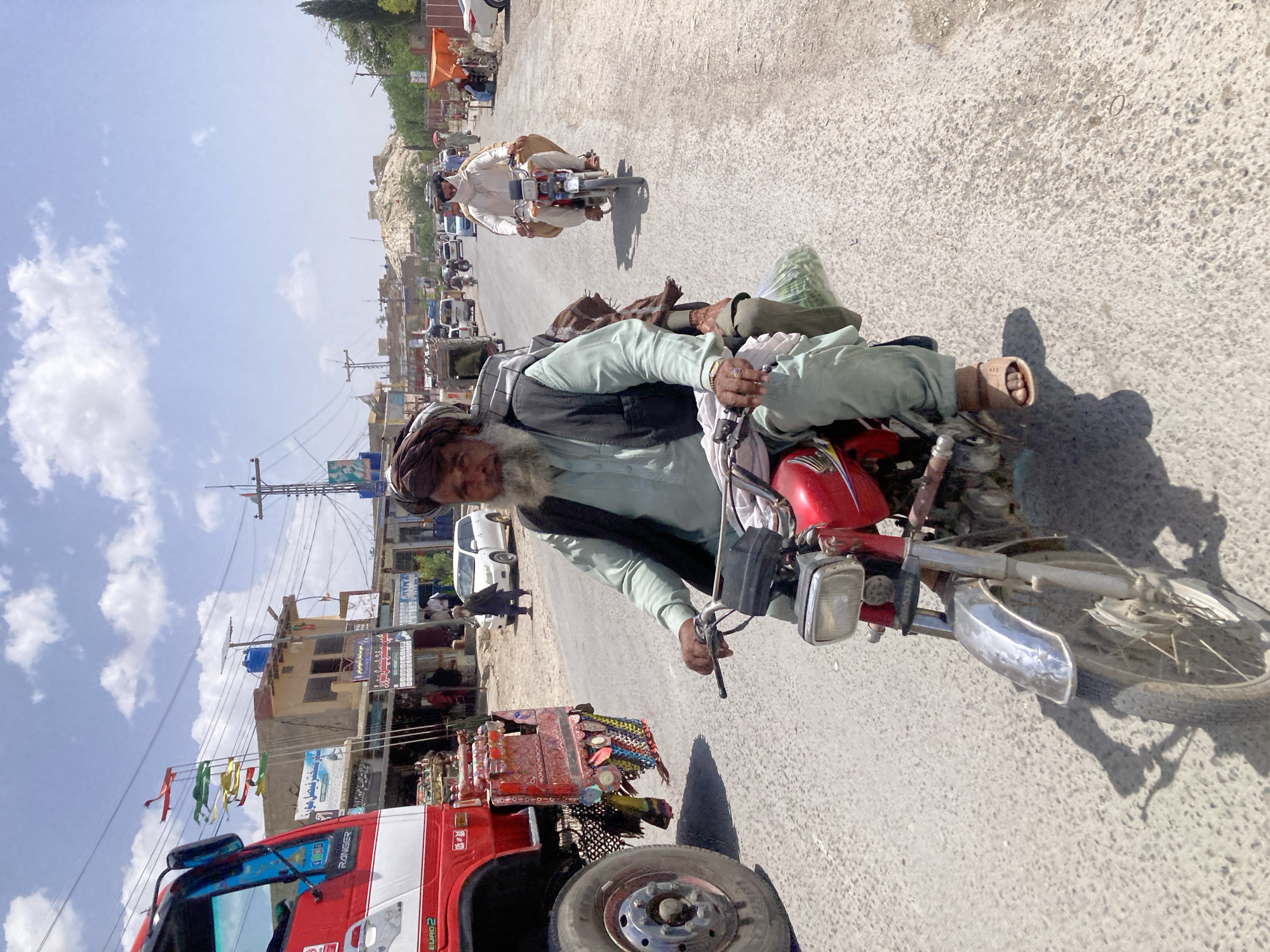 This image captures a bright, sunny street scene in Loralai, Pakistan, likely during the mid-morning or early afternoon given the strong shadows. The primary subject is an older man with a prominent white beard and a dark turban, wearing a light green shalwar kameez and a black vest. He is riding a red motorcycle, appearing focused on the road ahead. A green bag is visible on the motorcycle's seat behind him, and his right foot, clad in a sandal, rests near the engine.
The perspective is slightly elevated, possibly from another vehicle or a bystander standing on the roadside. The main road is paved with asphalt, showing some wear, while the edges feature dirt and rocky terrain.
In the middle ground, another man on a motorcycle, dressed in a light-colored traditional outfit, follows behind. Further down the road, numerous vehicles, including parked cars and possibly more motorcycles, line the street. The background reveals a bustling urban landscape with low-rise buildings housing shops and businesses, some with signs in local scripts (likely Urdu or Pashto) that are not clearly legible. Power lines and utility poles crisscross the sky, adding to the urban feel.
On the far left, a large red truck is visible, featuring distinct, colorful decorations on its rear, typical of regional truck art. Text "RANGER" and "EURO 2" can be seen on its side. The sky is a clear blue with scattered white clouds. In the distance, hills rise, suggesting the arid, mountainous geography of the region. The scene depicts a slice of daily life and transportation in Loralai, characterized by traditional attire, motorcycles, and local commerce.