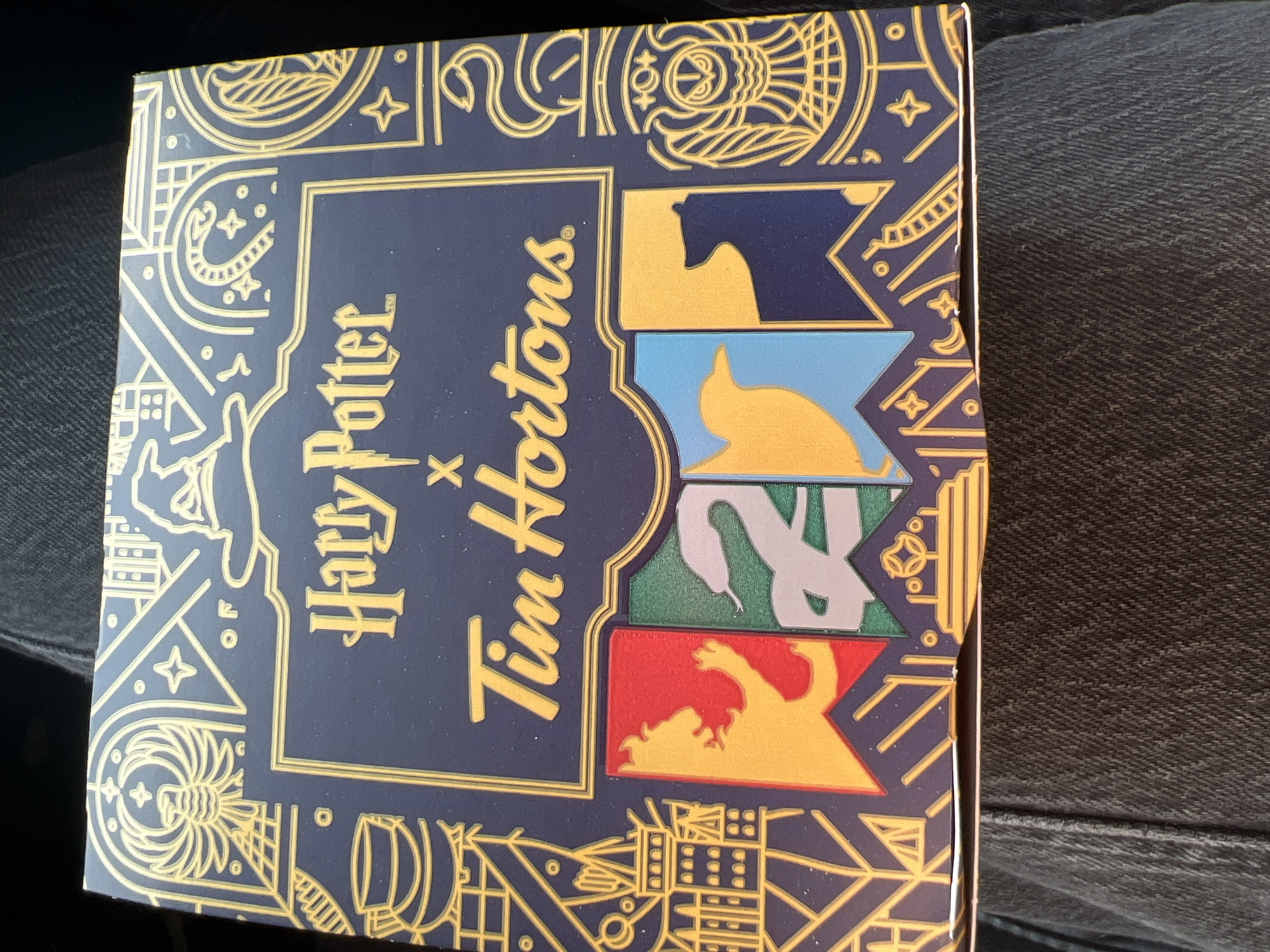 A close-up, top-down shot captures a dark blue and gold Harry Potter and Tim Hortons collaboration box. The box is decorated with ornate golden line art, featuring elements such as a Sorting Hat, a snake, an owl, and various magical symbols. Prominently displayed across the front of the box are the words "Harry Potter" and "Tim Hortons." Below these words, four rectangular banners are depicted, each with a different stylized crest: a lion on a red background, a serpent on a green background, a bird on a blue background, and a bear on a yellow background. The box rests on a person's lap, visible in the lower half of the image, wearing dark, textured denim jeans. The lighting suggests an indoor setting or an overcast day, with no discernible background beyond the person's clothing and the box. There is no information in the image to suggest a location in Revelstoke, Canada.