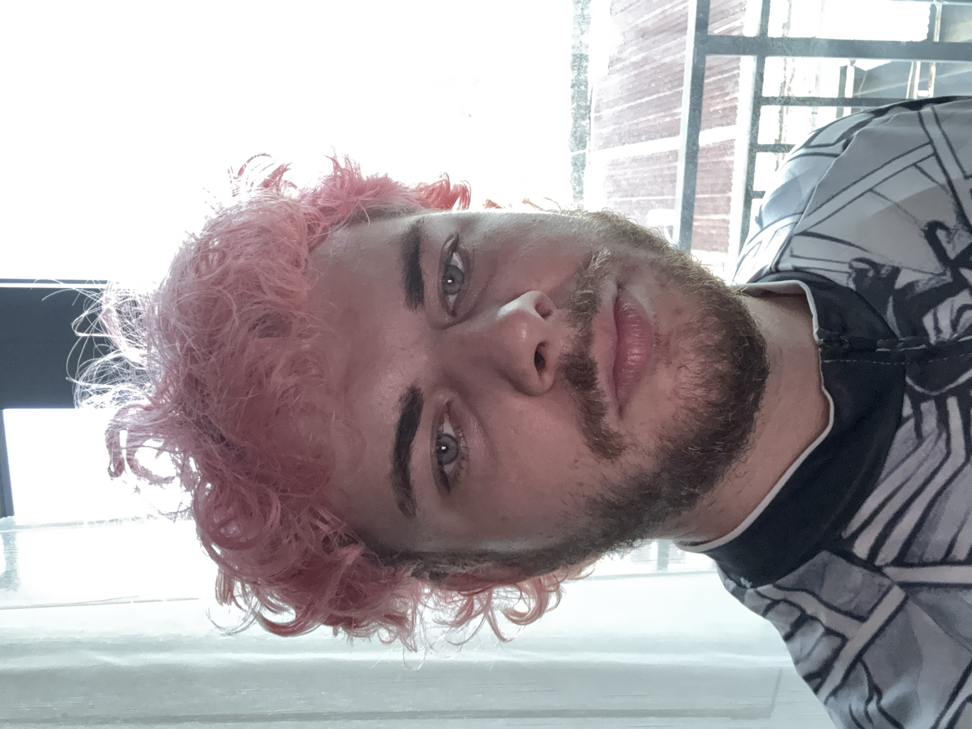 A close-up, tilted image shows a person with pink, curly hair and a beard. The person is wearing a shirt with a black and white geometric pattern. In the background, a window with metal bars is visible. The location and city of Đà Nẵng, Vietnam are not discernible from the image.