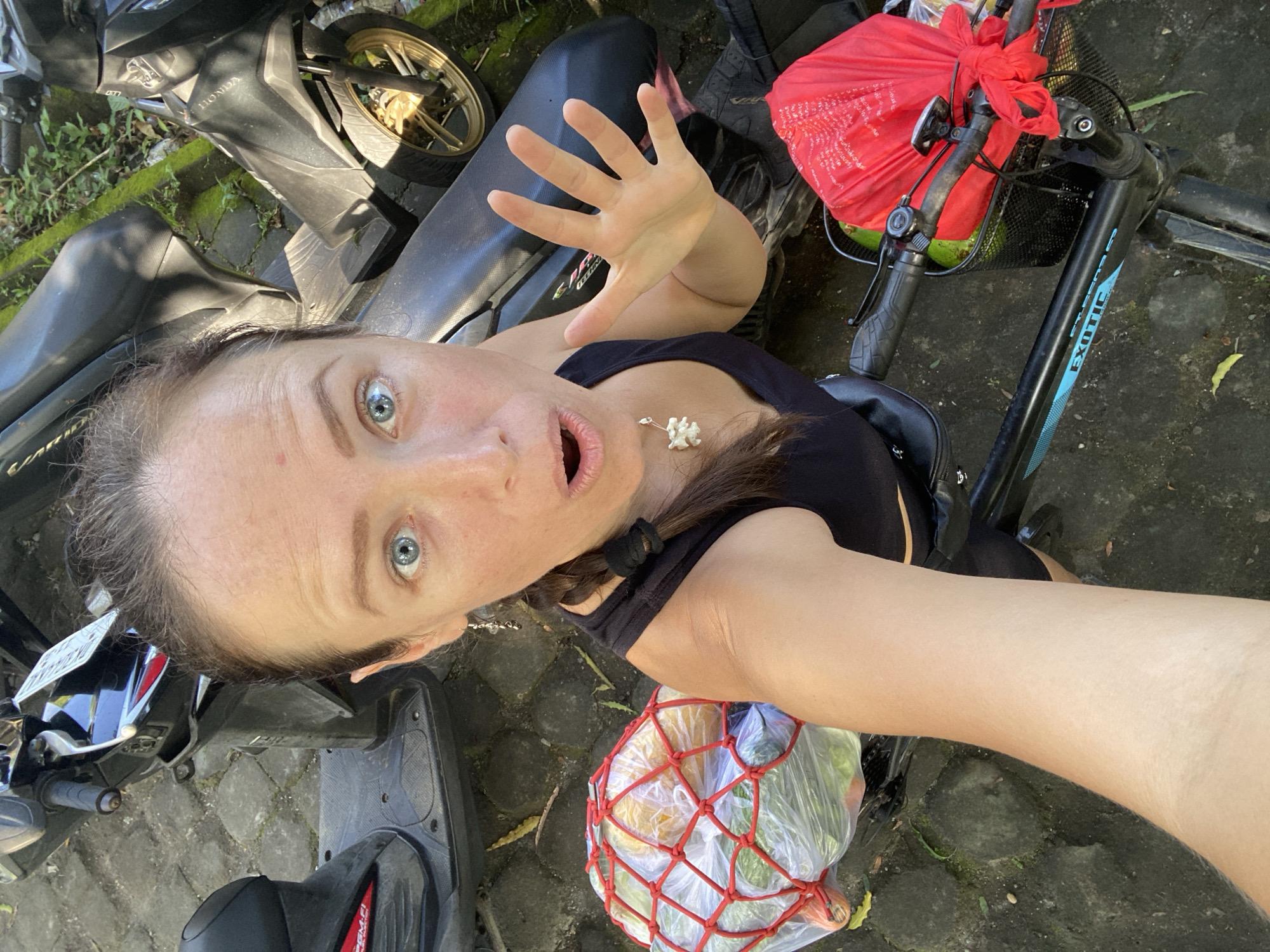 A fair-skinned woman with blue eyes is taking a selfie from a high angle. She has a surprised expression, with her mouth open and her left hand raised, palm facing forward. She's wearing a black top and a small, possibly floral, necklace. A portion of her brown hair is pulled back with a black hair tie.
To the left of the woman, two parked scooters are visible. The one closest to the viewer has a visible license plate. To the right, there's a bicycle with a front basket containing a red bag. Another net bag with produce is near her body. The location appears to be outdoors, on a paved surface with moss growing between the stones. Based on the prompt, this is occurring in Sayan, Indonesia.