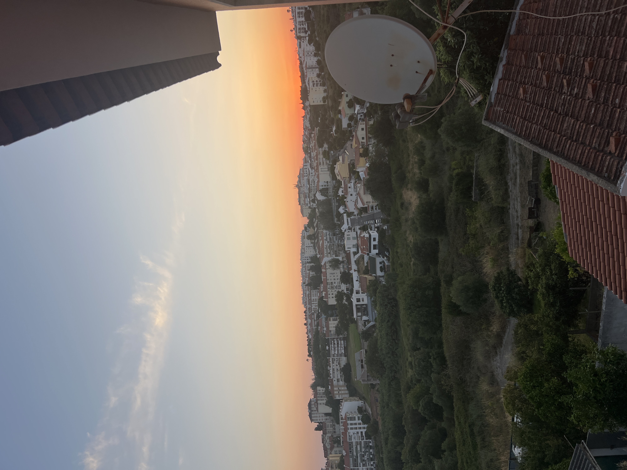 The image depicts a panoramic view of a residential area in Cascais, Portugal, during what appears to be sunset. The sky transitions from a clear pale blue at the top to vibrant shades of orange and yellow near the horizon, indicating the sun is setting or has recently set. A wispy cloud formation is visible in the upper left quadrant.
In the foreground, the tiled roof of a building is prominent, with a portion of a wall and overhang visible on the right side. A white satellite dish, mounted on a rusty pole with several wires connected, is positioned in the mid-ground, slightly to the right of the center.
Beyond the foreground structures, the landscape slopes downwards and then upwards, covered with dense green vegetation and trees. Scattered throughout this verdant area and on the rising hills are numerous residential buildings, mostly low to mid-rise apartments and houses, characterized by white, cream, and red tiled roofs. The buildings are densely packed in some areas, forming a town or city sprawl. Some streets and individual vehicles are discernible within the built-up areas. The overall environment suggests an urban or suburban setting with a significant amount of green space interspersed with residential development. No people or animals are visible in the image.