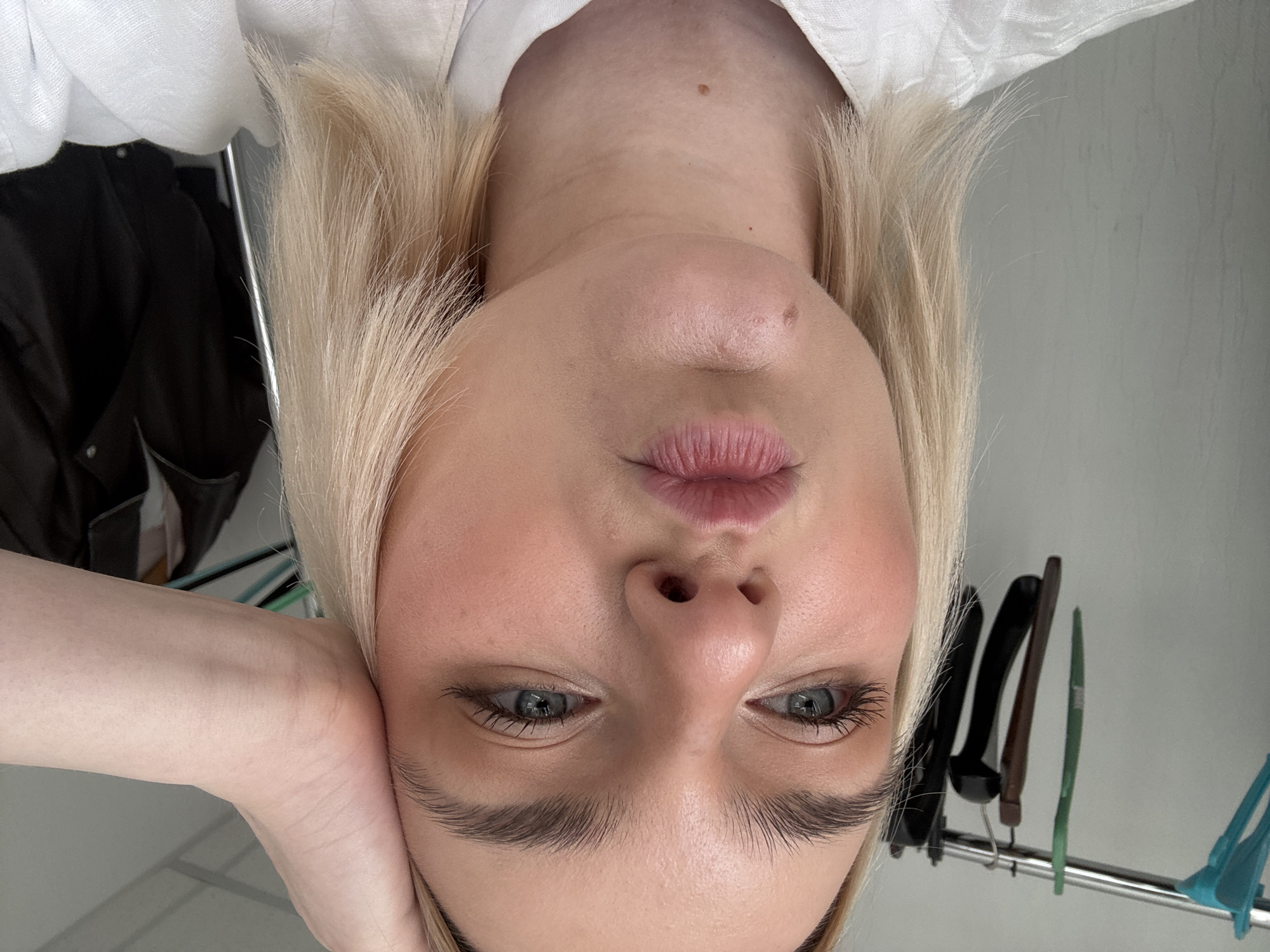 A close-up, upside-down selfie of a fair-skinned woman with blonde hair. Her face fills the majority of the frame. She is resting her left cheek on her hand. The background shows clothing hanging on a rack and a white wall. The location is not discernible from the image; no clues indicate Saint Petersburg, Russia.